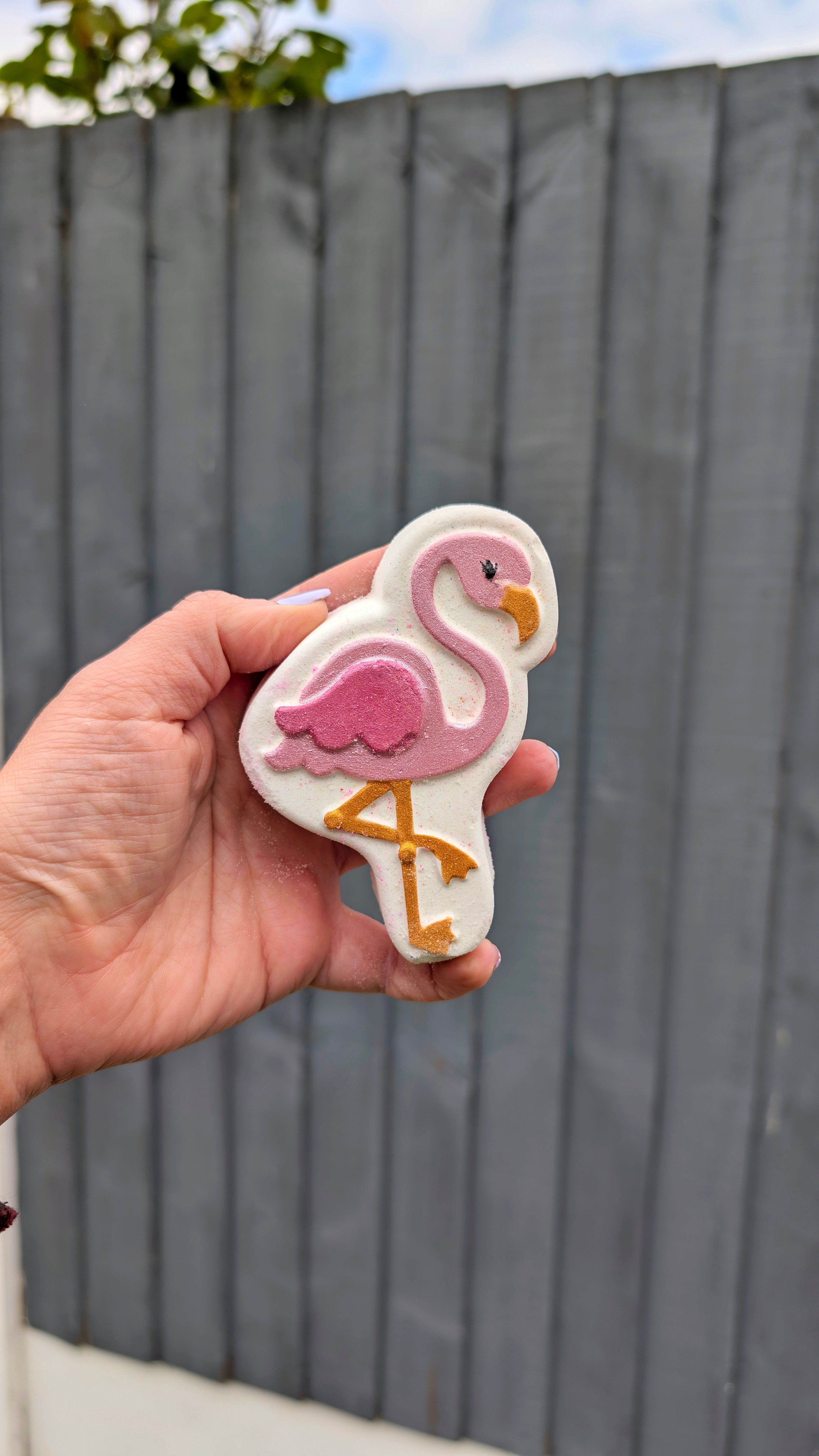 Hand holding a flamingo shaped bath bomb from Fizz Bomb Cosmetics, featuring pink, white, and gold detailing – a fun, tropical themed bath treat perfect for gifting or relaxing self care days.
