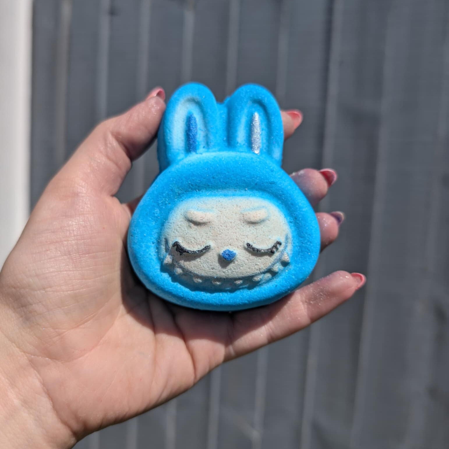 Hand holding a blue labubu face shaped bath bomb, with a grey fence in the background