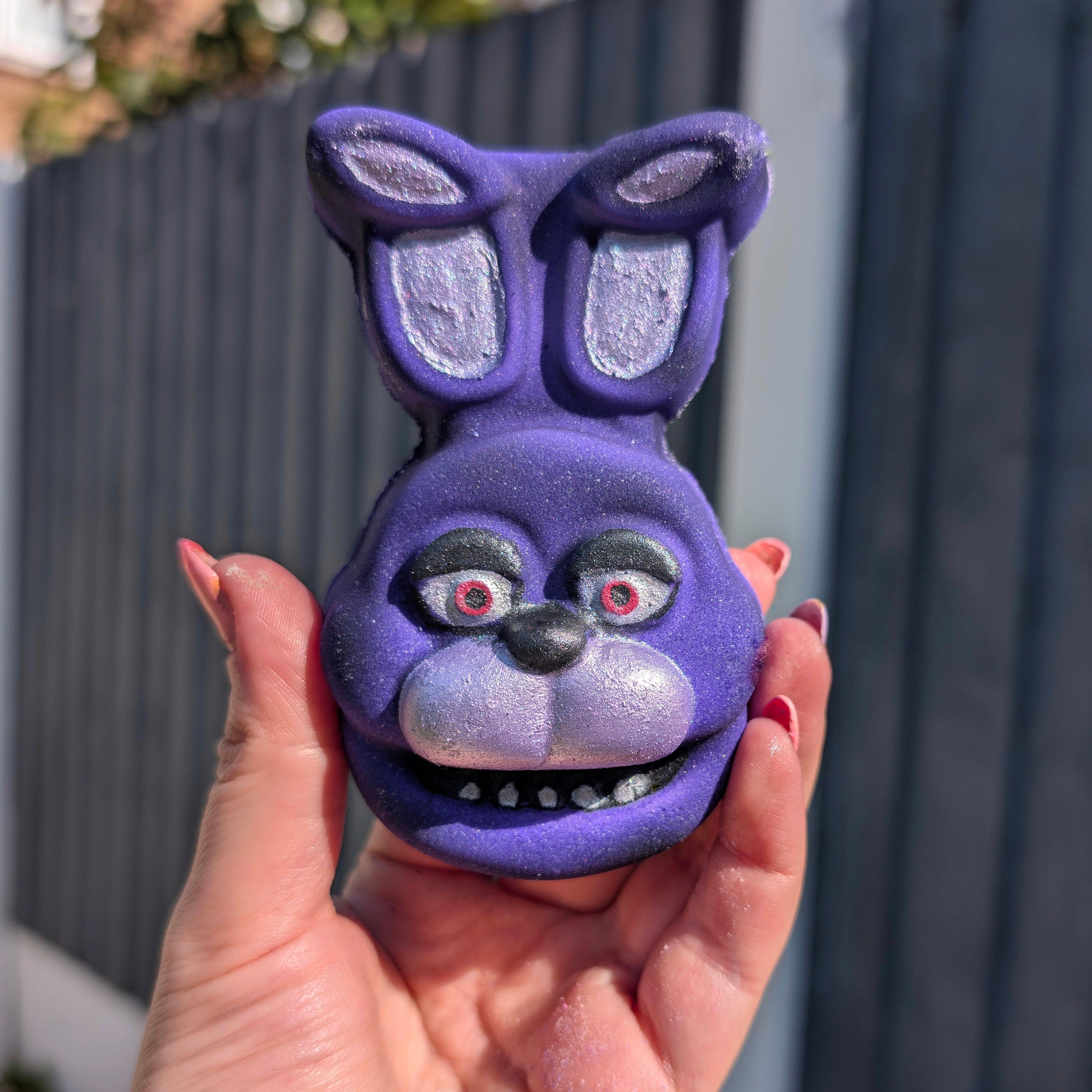 Purple Bonnie FNAF character head with bunny ears held in a hand against a grey fence background
