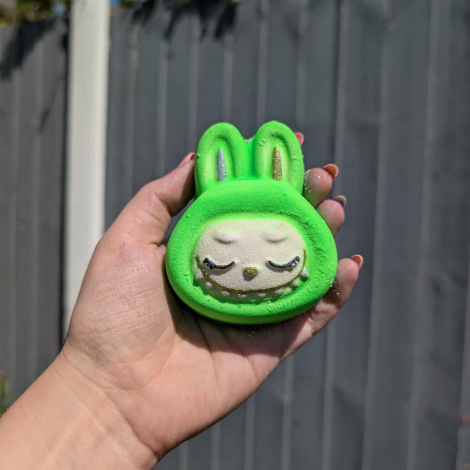 Hand holding a green labubu face shaped bath bomb, with a grey fence in the background
