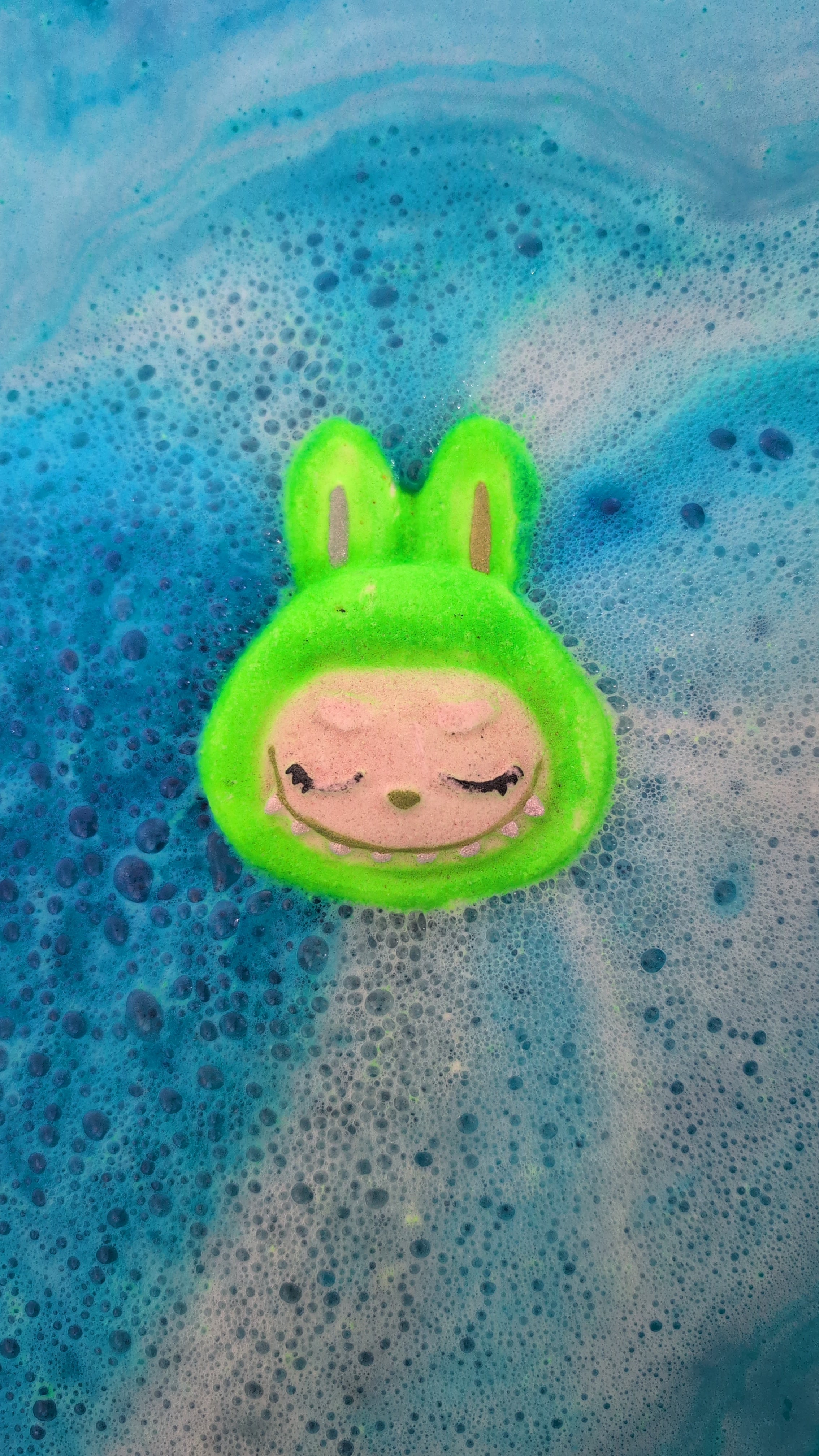 Green labubu face shaped bath bomb, in use in the bath, with blue colour bursting out.
