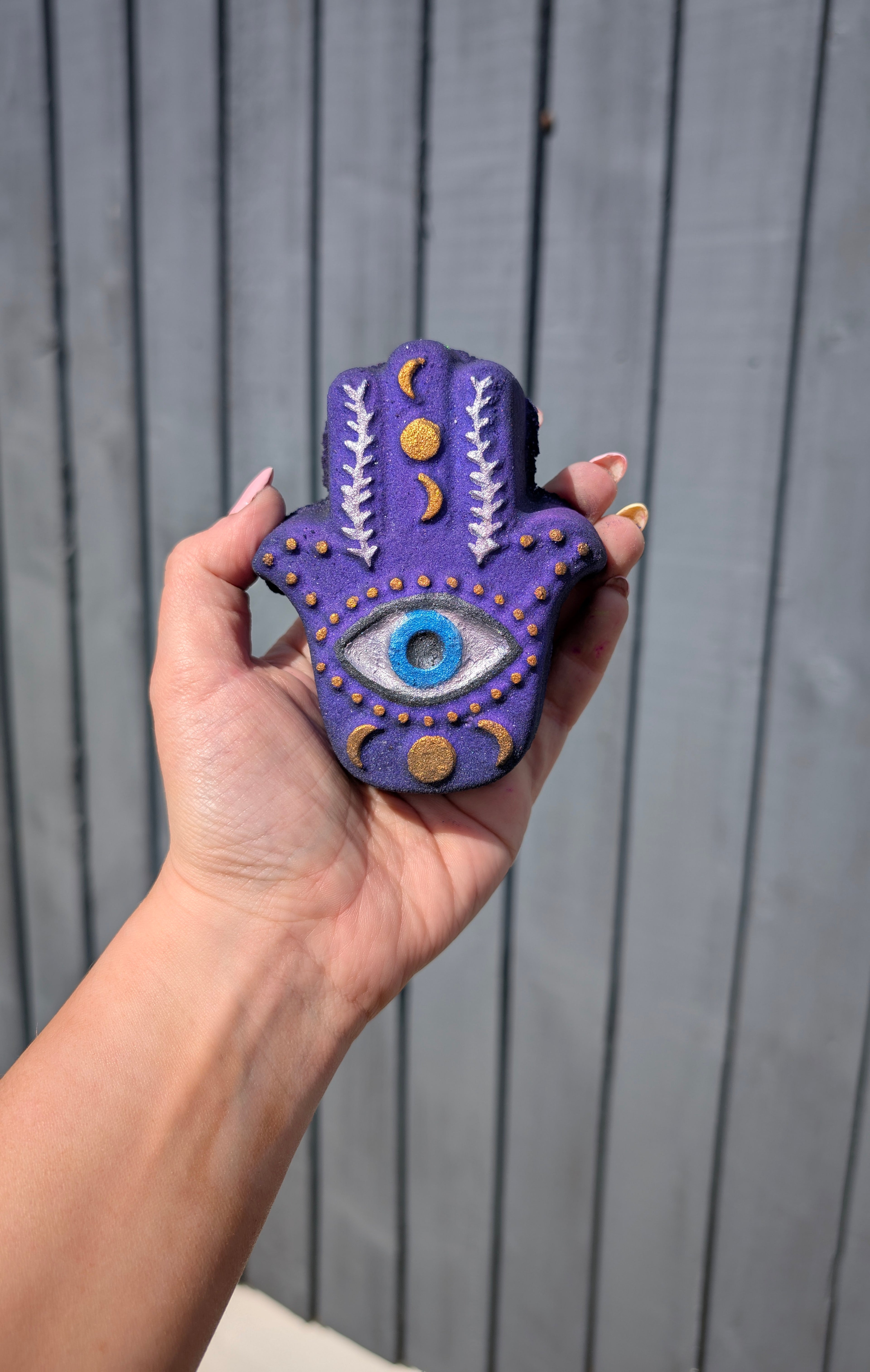 Hand holding a hamsa hand shaped bath bomb from Fizz Bomb Cosmetics, featuring purple, white, and gold detailing – a fun, mystical themed bath treat perfect for gifting or relaxing self care days.