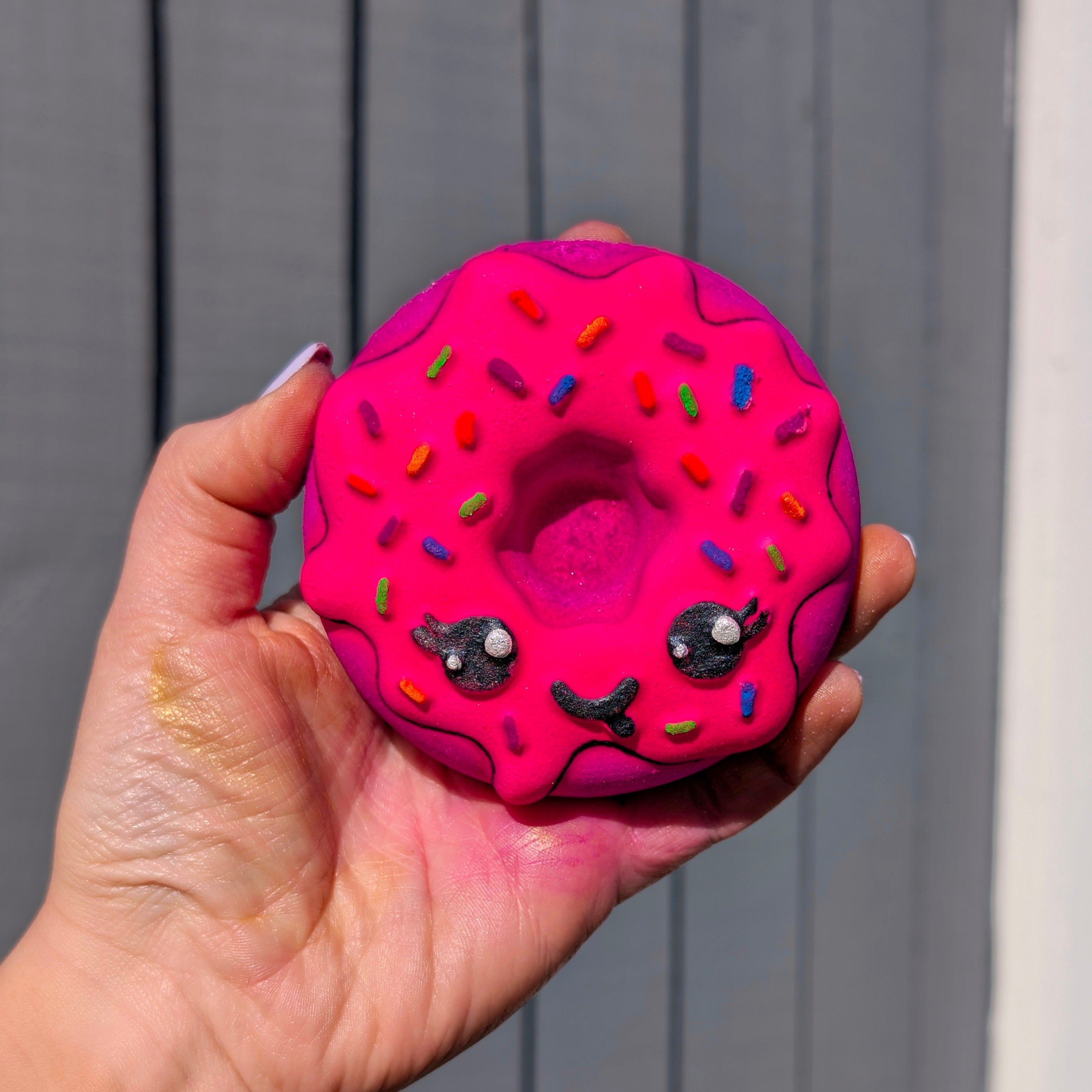 Hand holding a kawaii doughnut shaped bath bomb from Fizz Bomb Cosmetics, featuring pink,black and popping colours detailing – a fun, cake themed bath treat perfect for gifting or relaxing self care days.