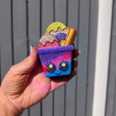 Colorful Kawaii ice cream shaped bath bomb held by a hand with a gray fence and blue sky in the background