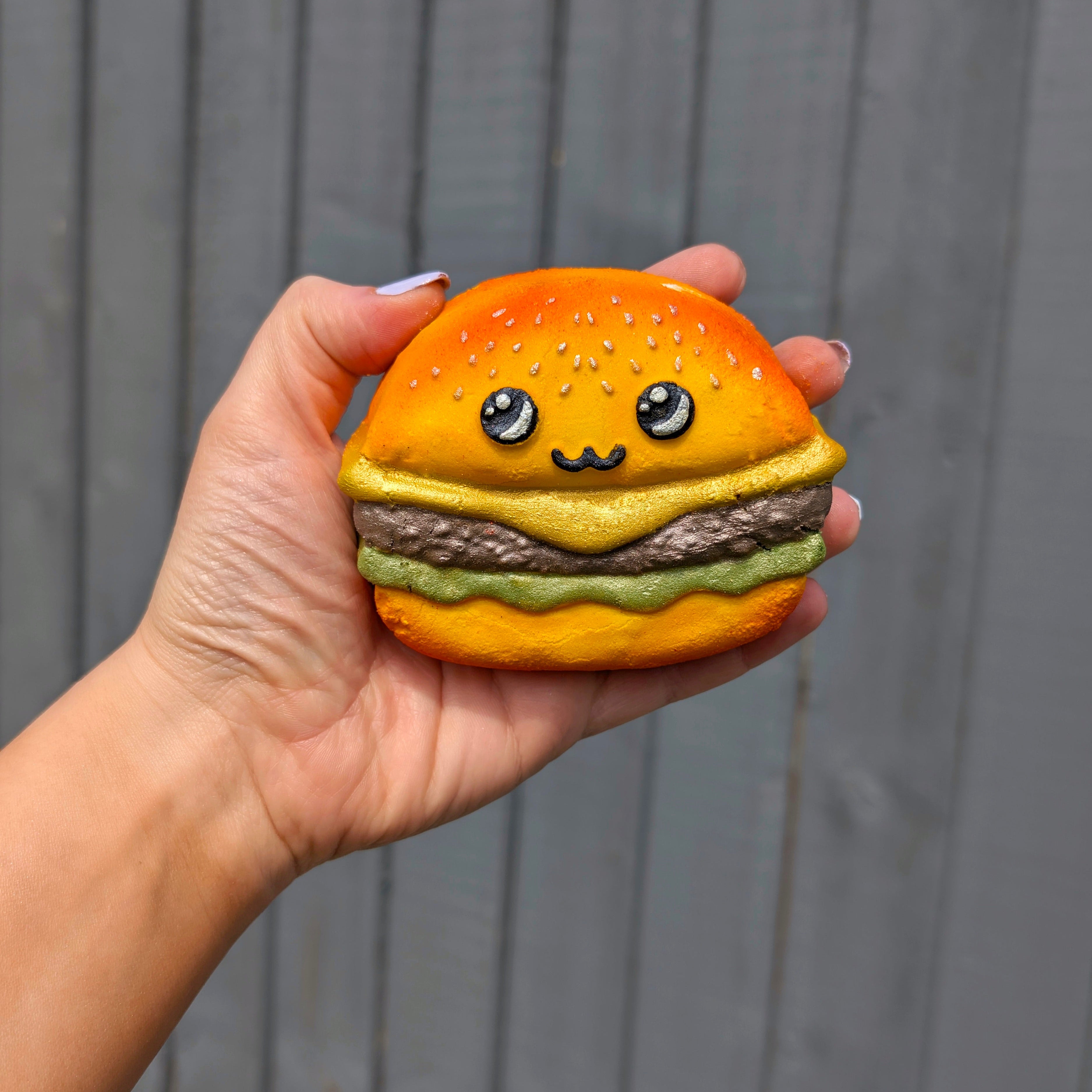 Hand holding a kawaii burger shaped bath bomb from Fizz Bomb Cosmetics, featuring yellow, green and white detailing – a fun, burger themed bath treat perfect for gifting or relaxing self care days.