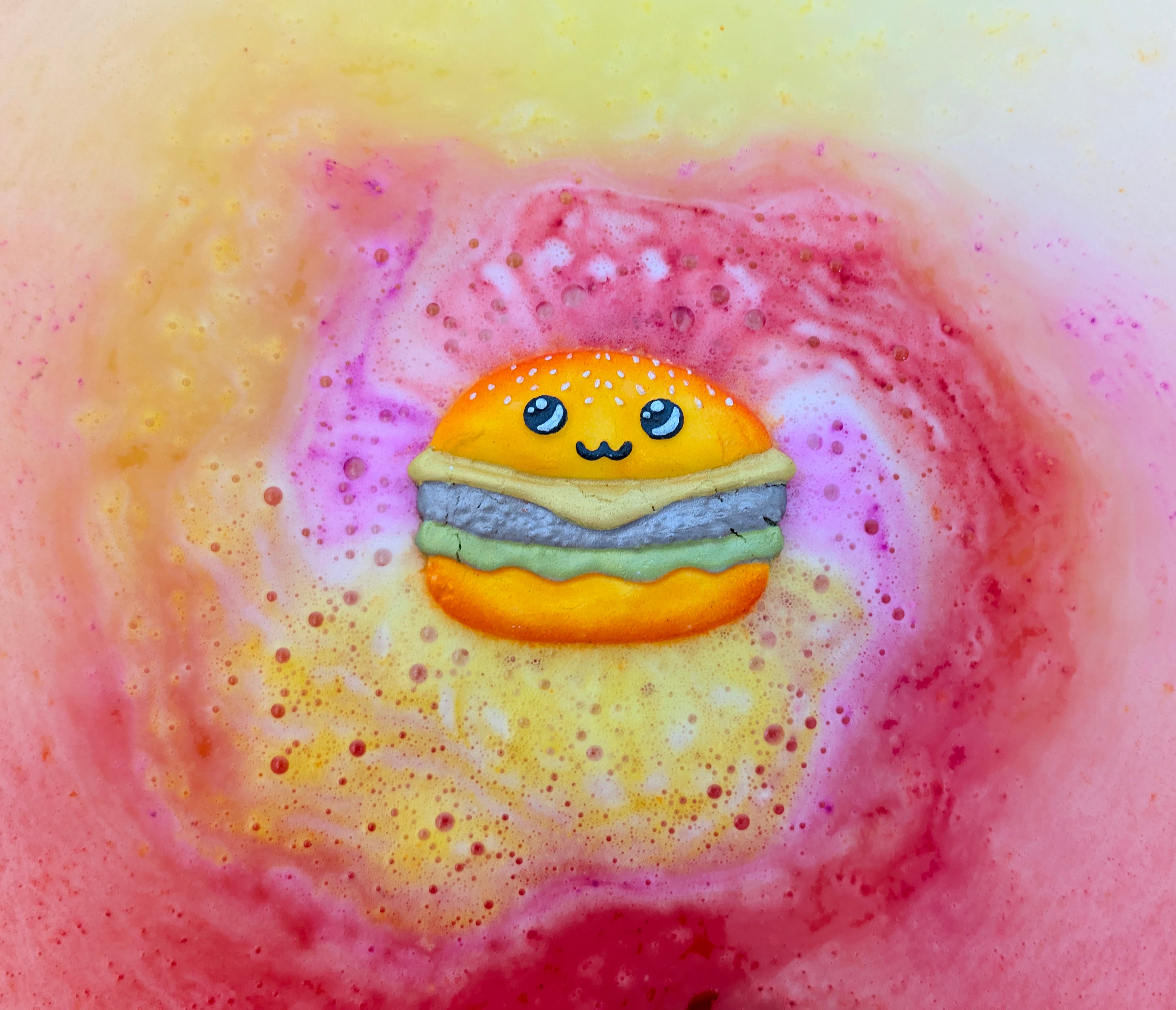 Kawaii burger shaped bath bomb from Fizz Bomb Cosmetics in use in the bath, featuring pink and yellow colours bursting out into the water – a fun, burger themed bath treat perfect for gifting or relaxing self care days.