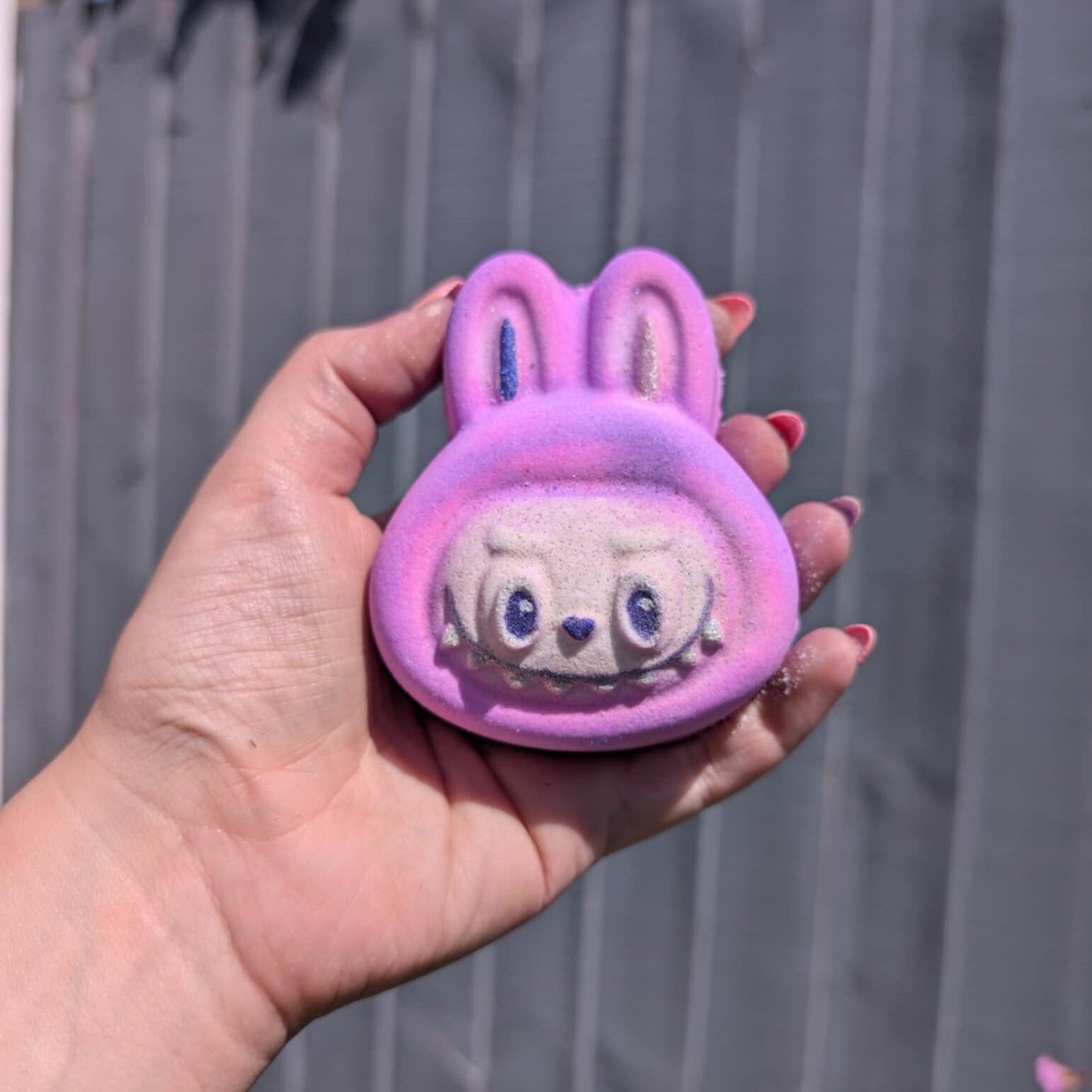 Hand holding pink and purple shaded labubu bath bomb, with grey fence in the background.