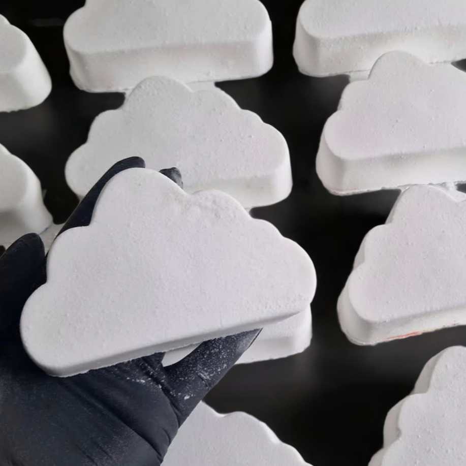 A hand holding a bath bomb shaped like a cloud, with a background of similar bath bombs arranged on a surface.