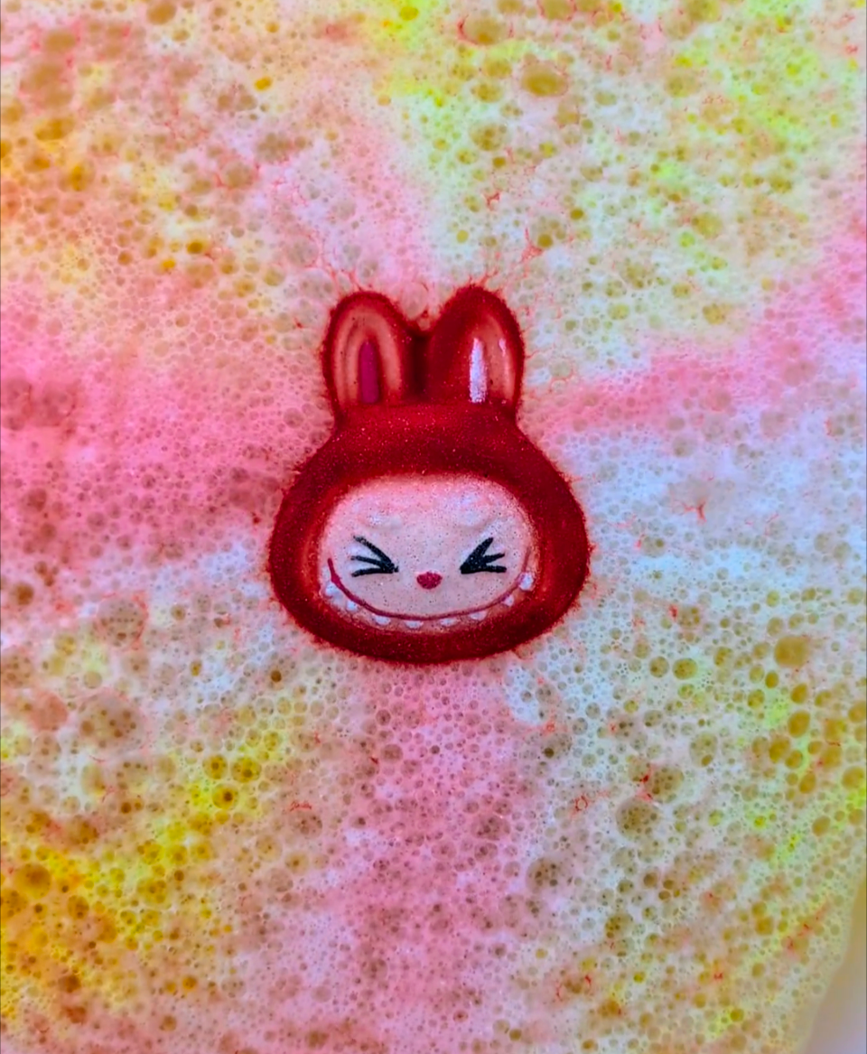 Red labubu face shaped bath bomb, in use in the bath, with coloursred and yellow colours bursting out.
