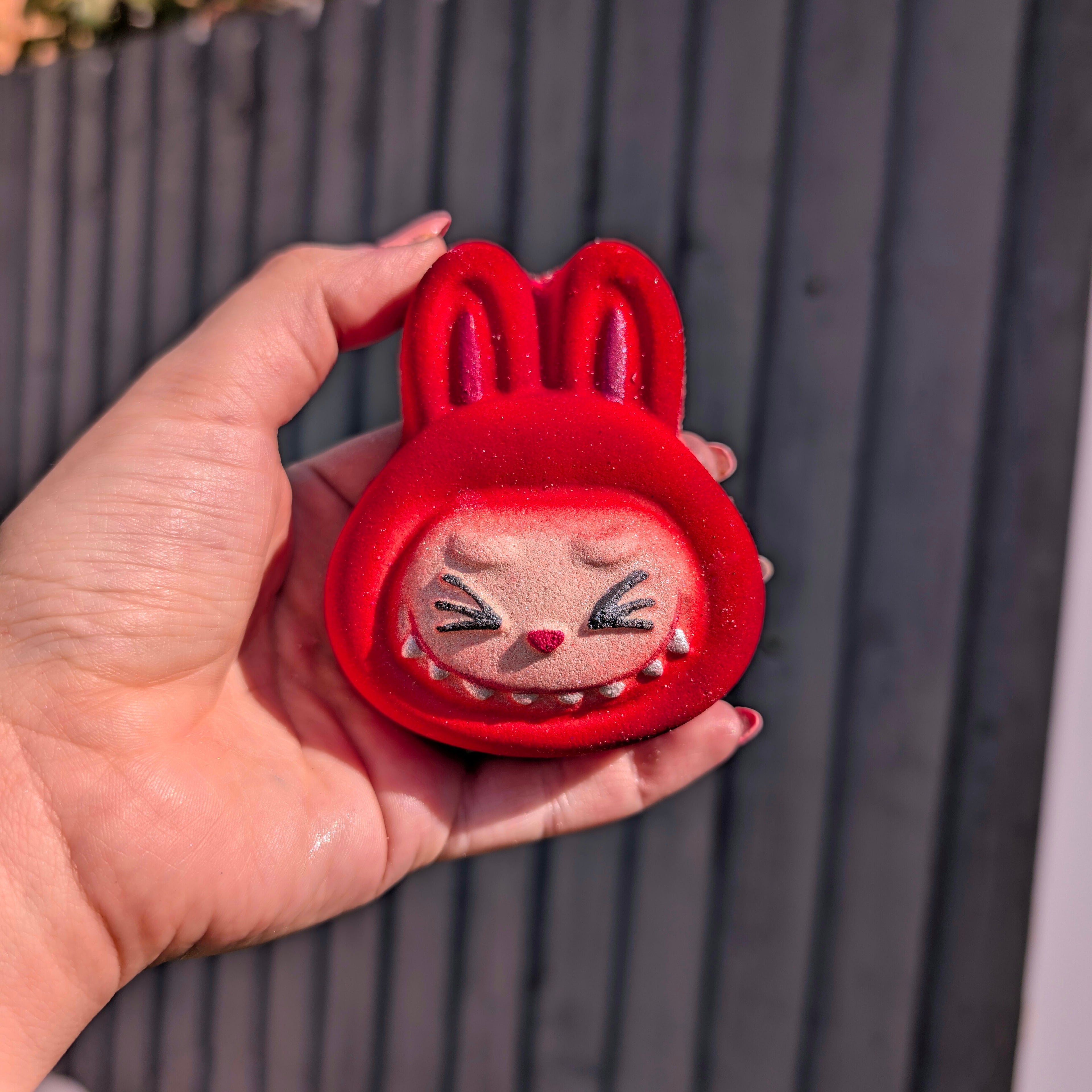 Hand holding a red labubu shaped face bath bomb, with a grey fence in the background.