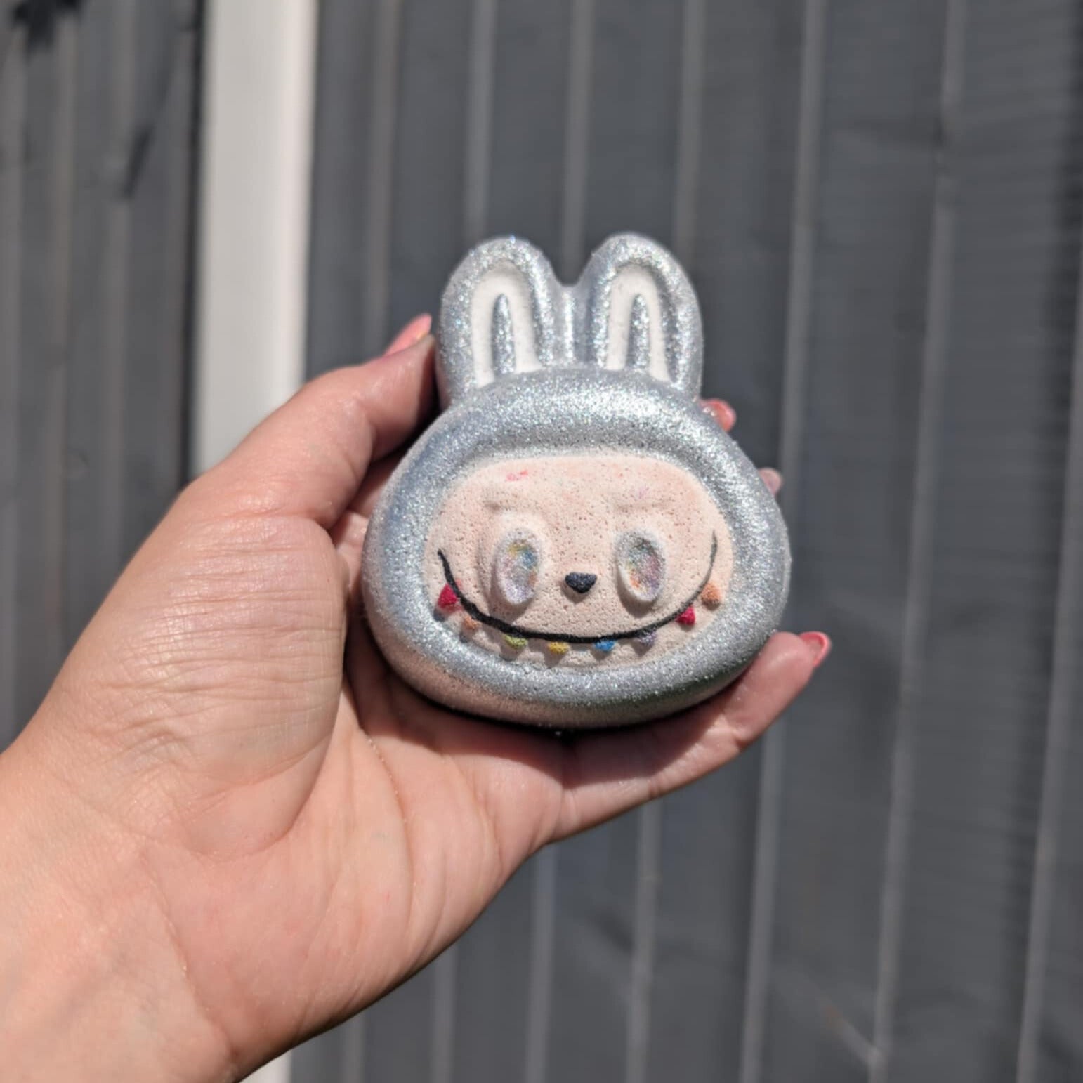 Hand holding silver labubu face bath bomb with a grey fence in the background