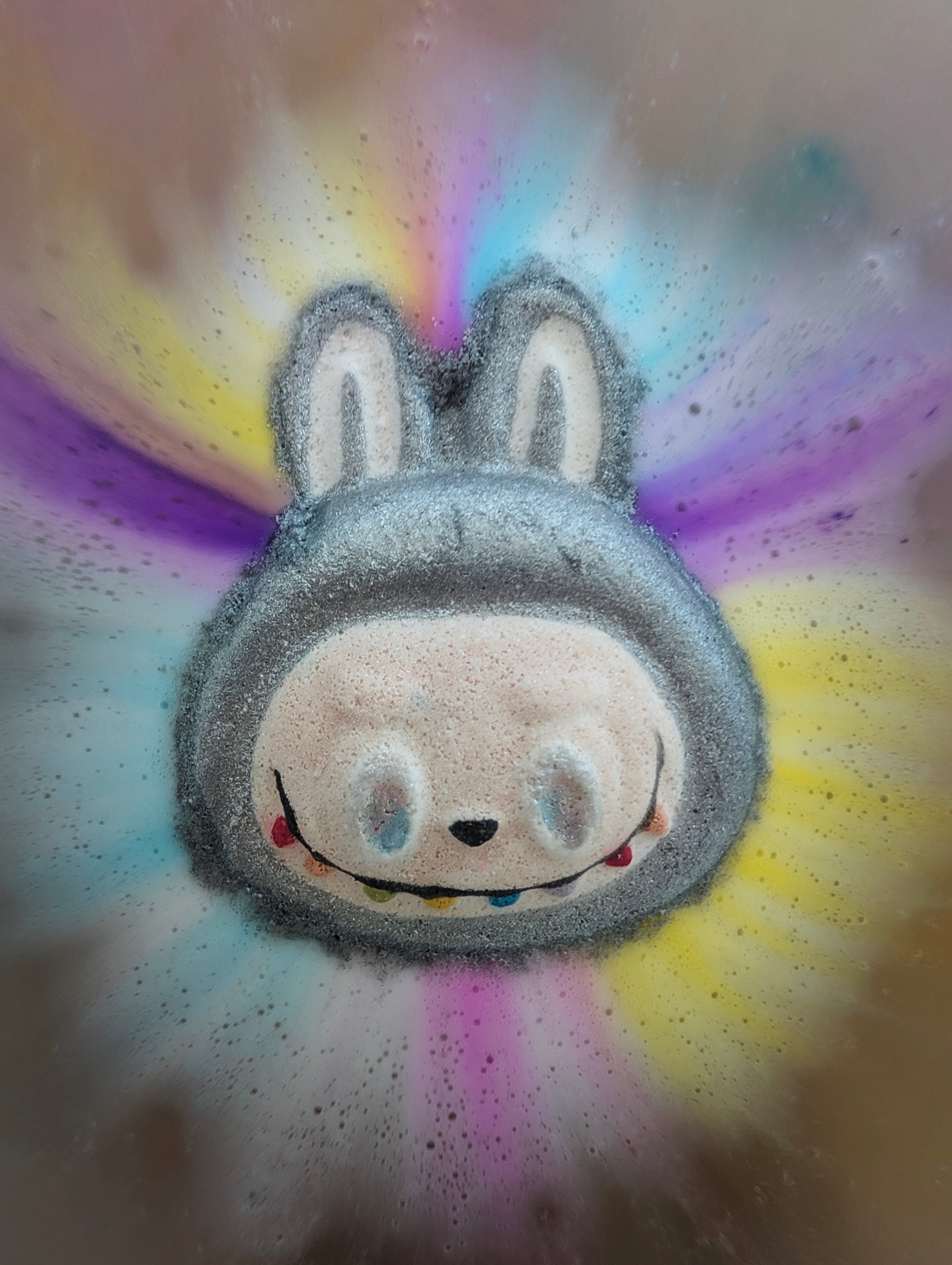 Silver labubu face shaped bath bomb, in use in the bath, with colours purple, blue, pink and yellow colours bursting out.