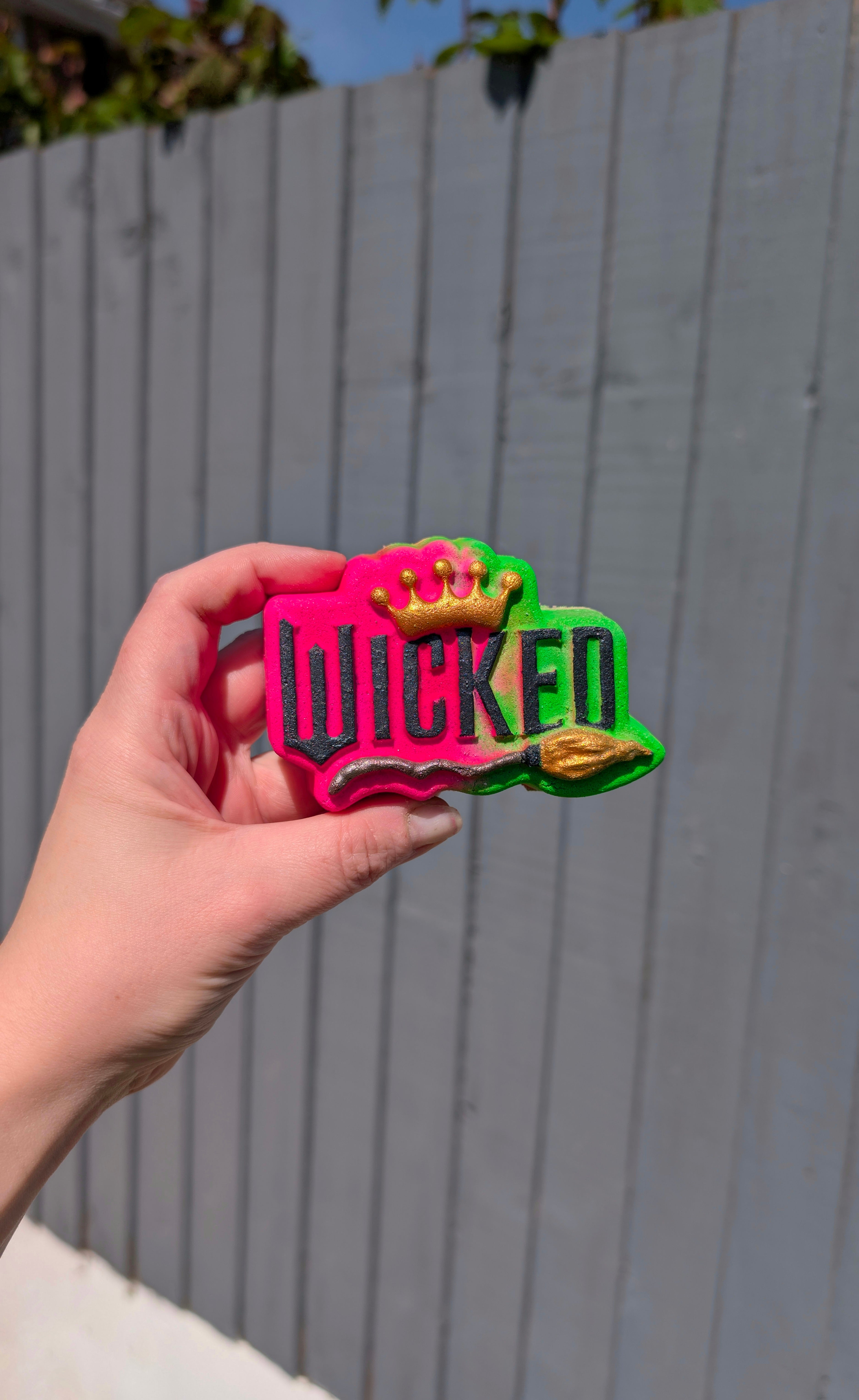 Hand holding a wicked shaped bath bomb from Fizz Bomb Cosmetics, featuring bright pink, green, black and gold detailing – a fun, wicked themed bath treat perfect for gifting or relaxing self care days.