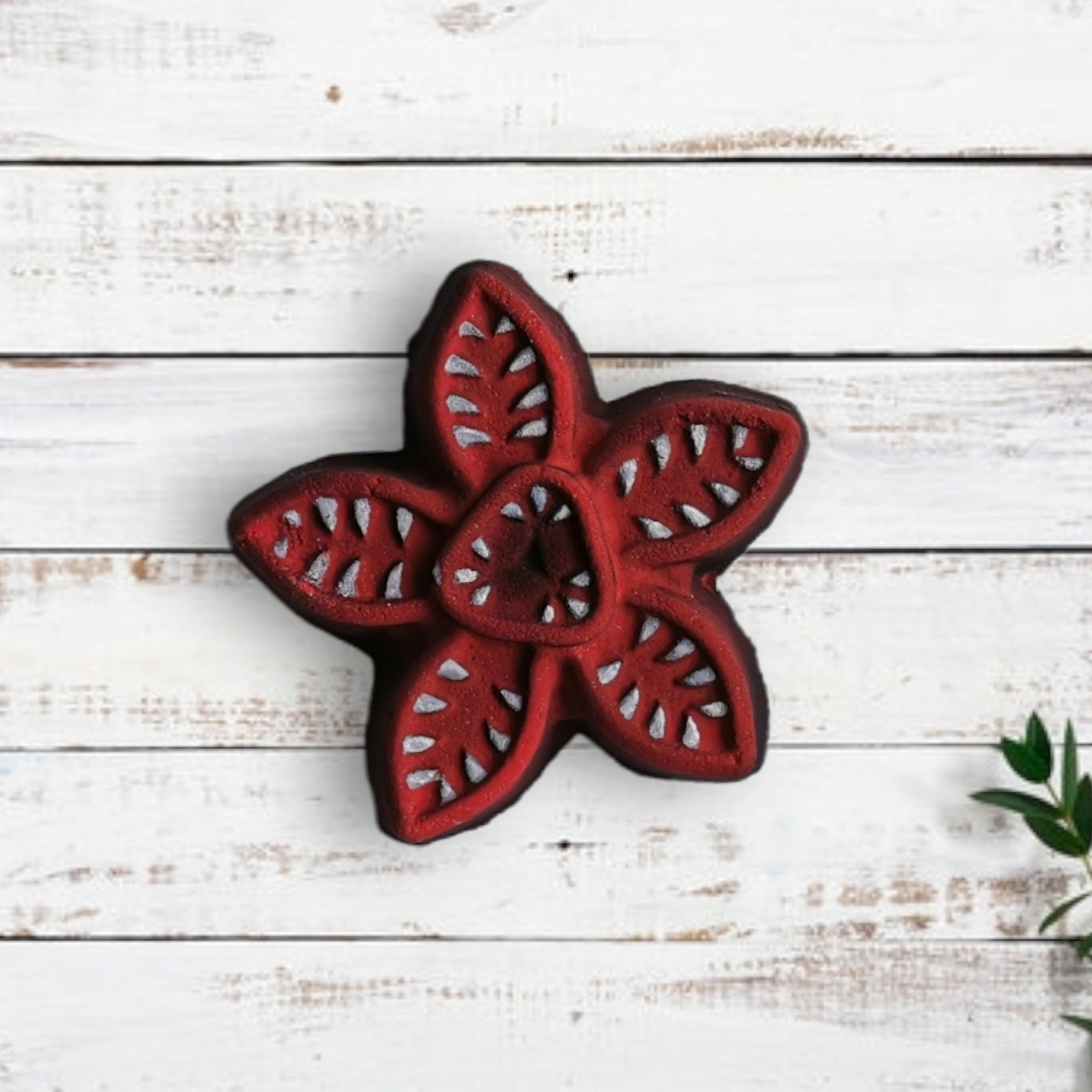 A red, stranger things demogorgon shaped bath bomb with a geometric pattern, placed on a white wooden surface with green leaves partially visible.