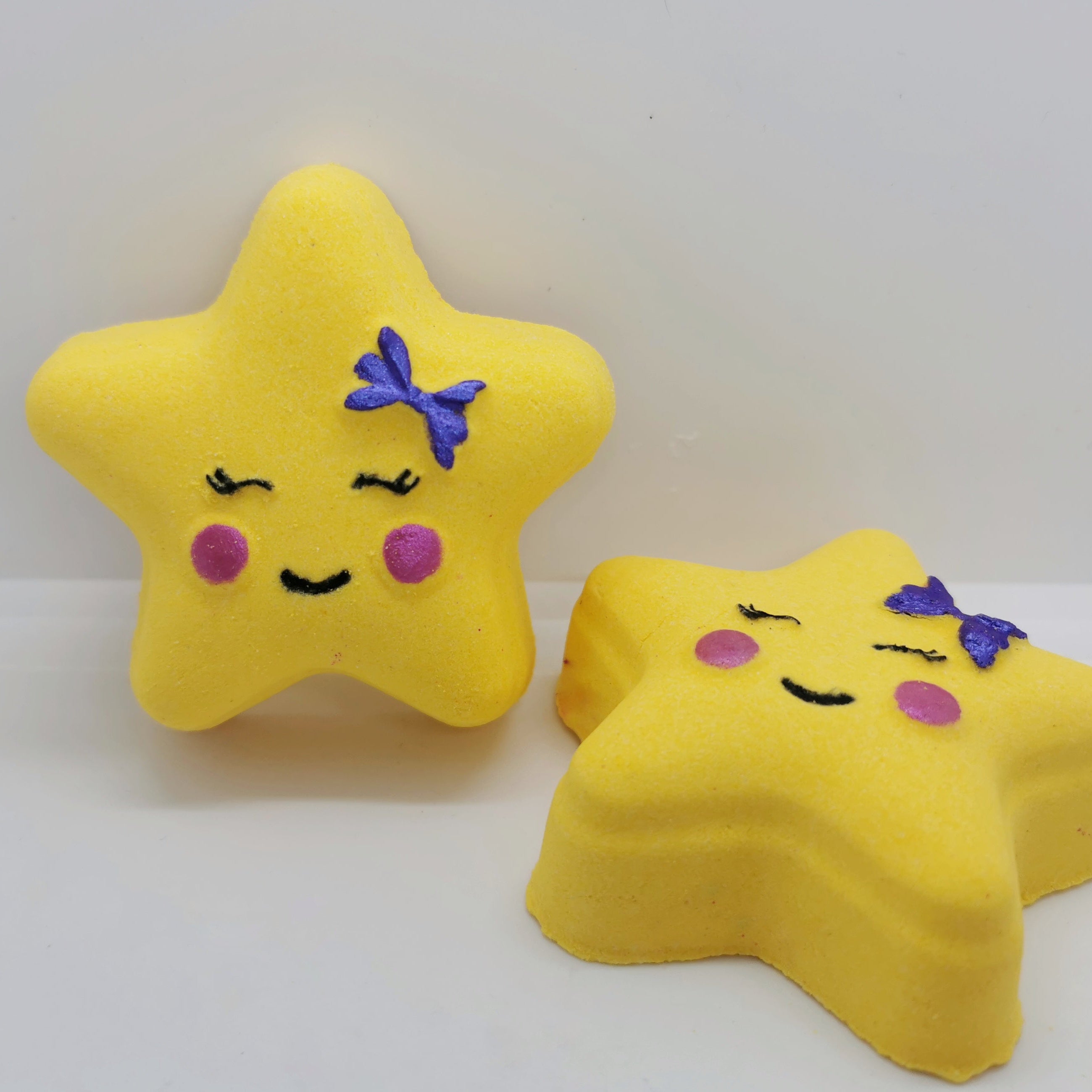 Two yellow bath bombs shaped like stars with simplistic facial features and a purple bow on each. One has a pink blush and the other has a purple blush.