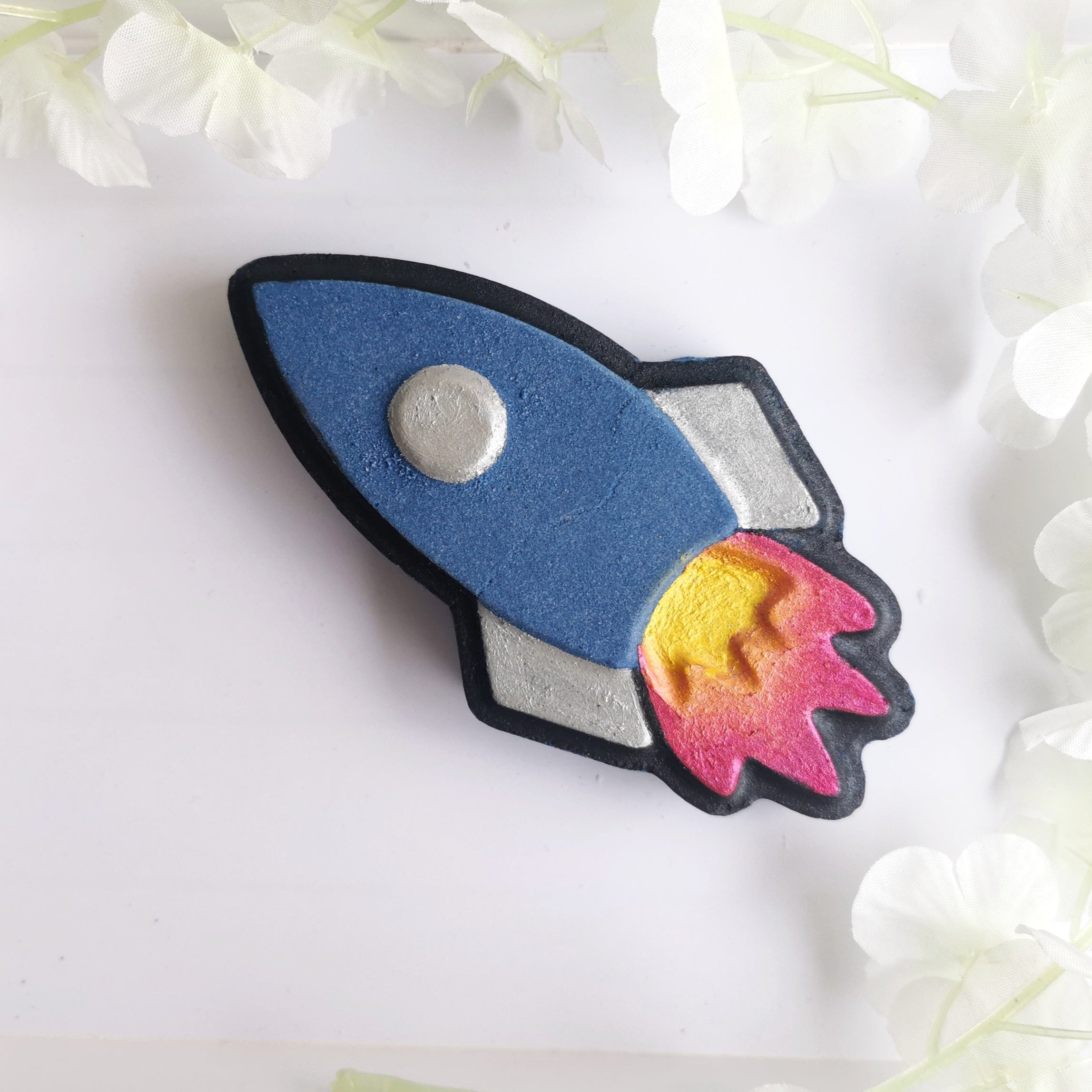 A bath bomb shaped like a rocket with a blue body, red and yellow fiery trails, and a grey nose cone, placed on a background with white floral elements.