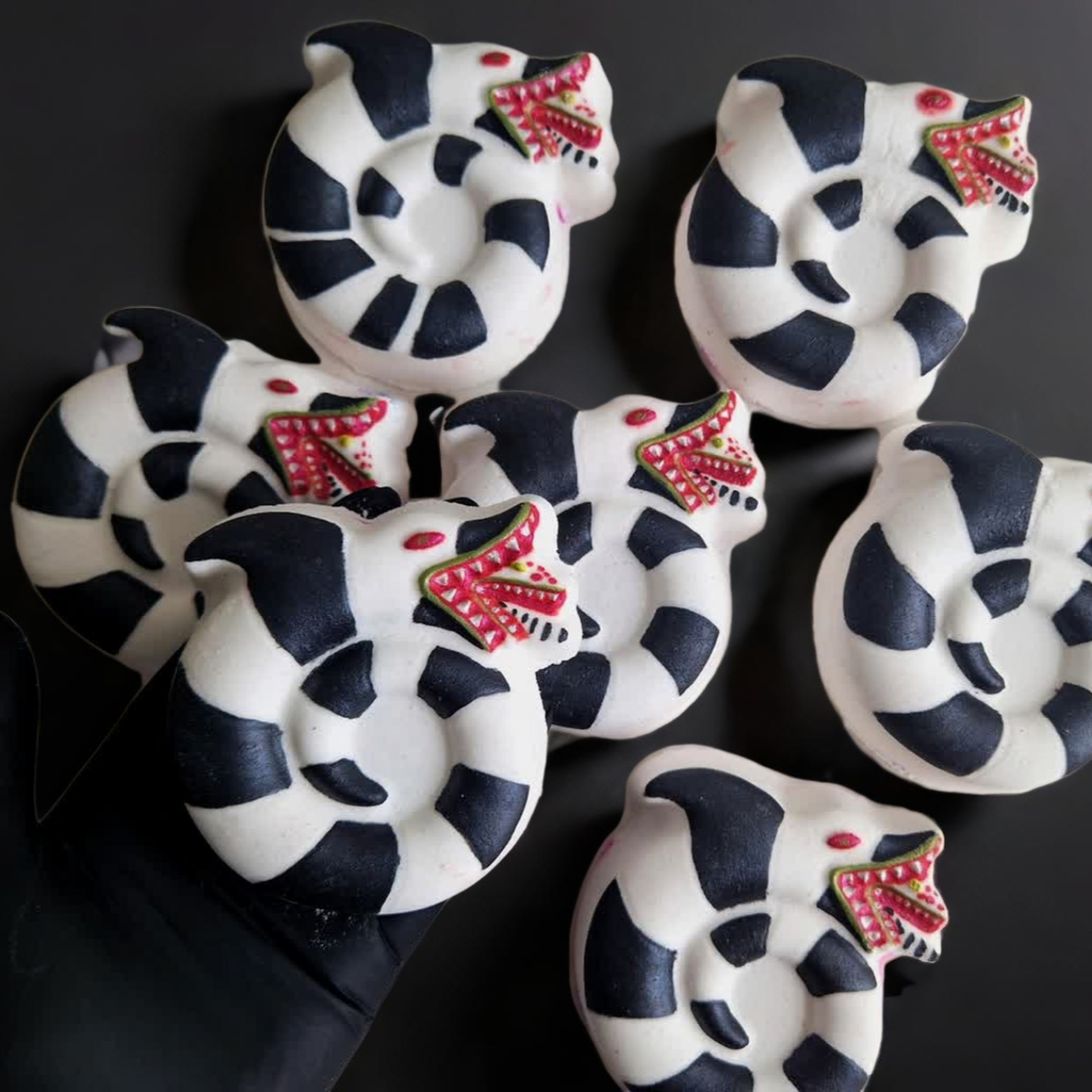 Hand holding a sandworm shaped bath bomb from Fizz Bomb Cosmetics, on a black background. Featuring red and black detailing – a fun, beetlejuice themed bath treat perfect for gifting or relaxing self-care days.