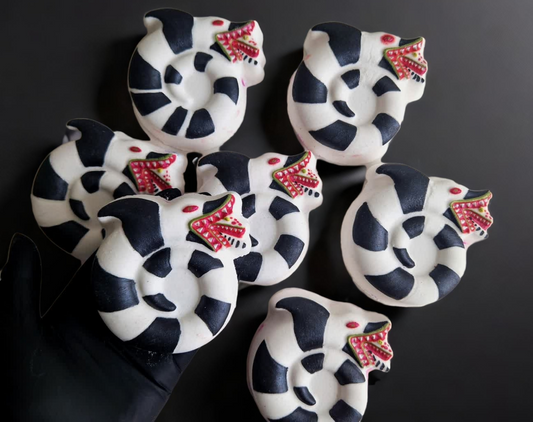 Hand holding a sandworm shaped bath bomb from Fizz Bomb Cosmetics, on a black background. Featuring red and black detailing – a fun, beetlejuice themed bath treat perfect for gifting or relaxing self-care days.