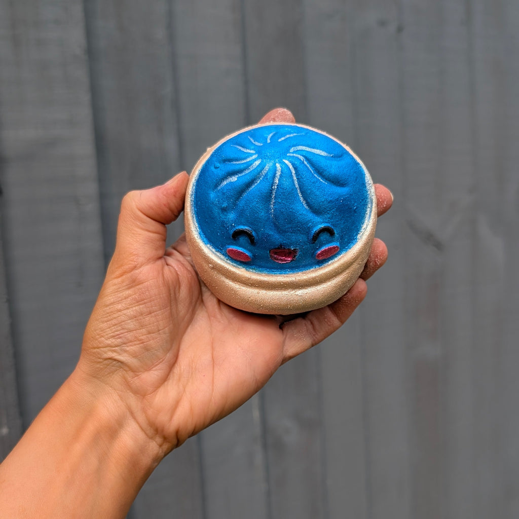 A hand holding a round dumpling bath bomb with a cute smiling face, set against a dark grey wooden plank background.