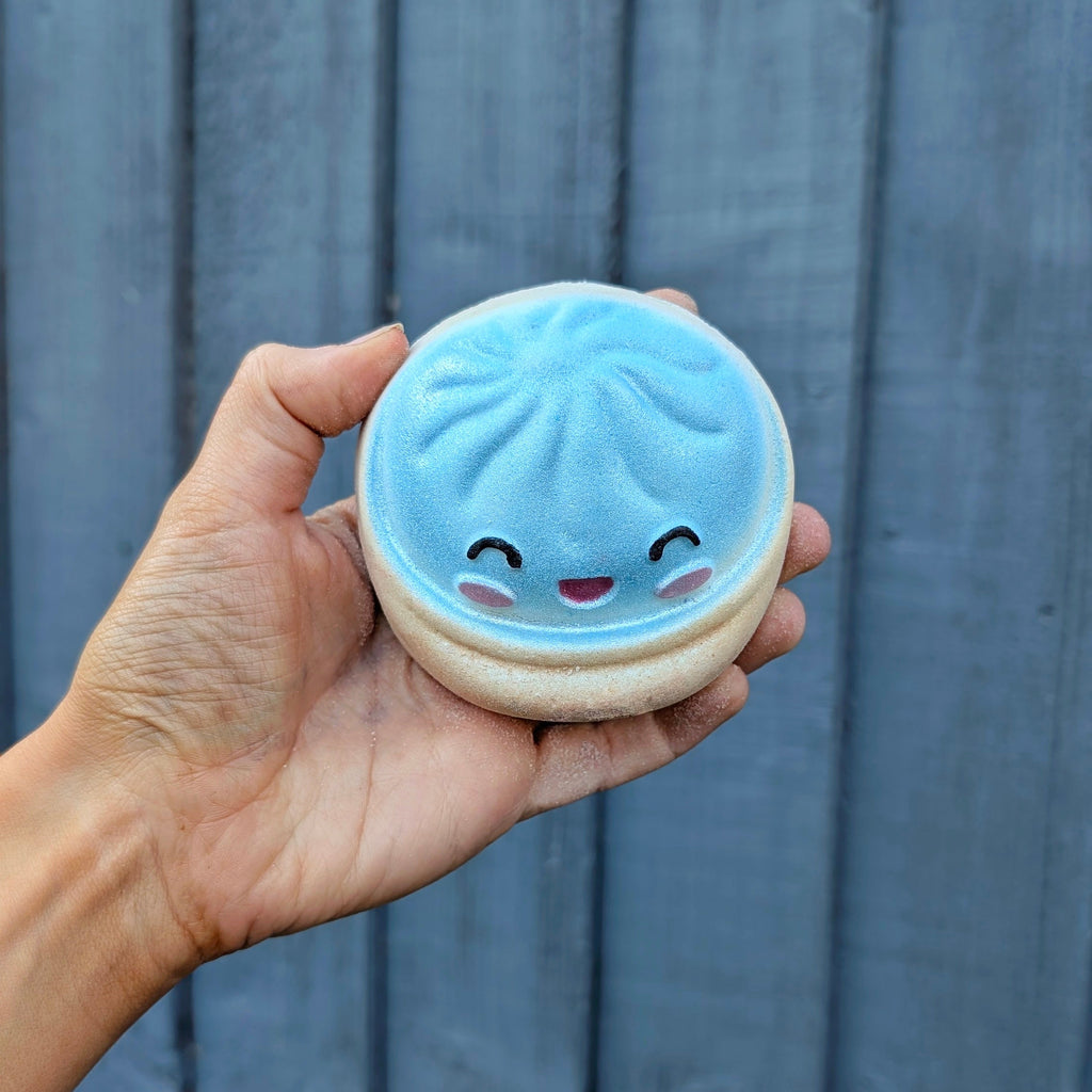A hand holding a small round, light blue and beige dumpling bath bomb with a cute smiling face, set against a dark grey wooden plank background.