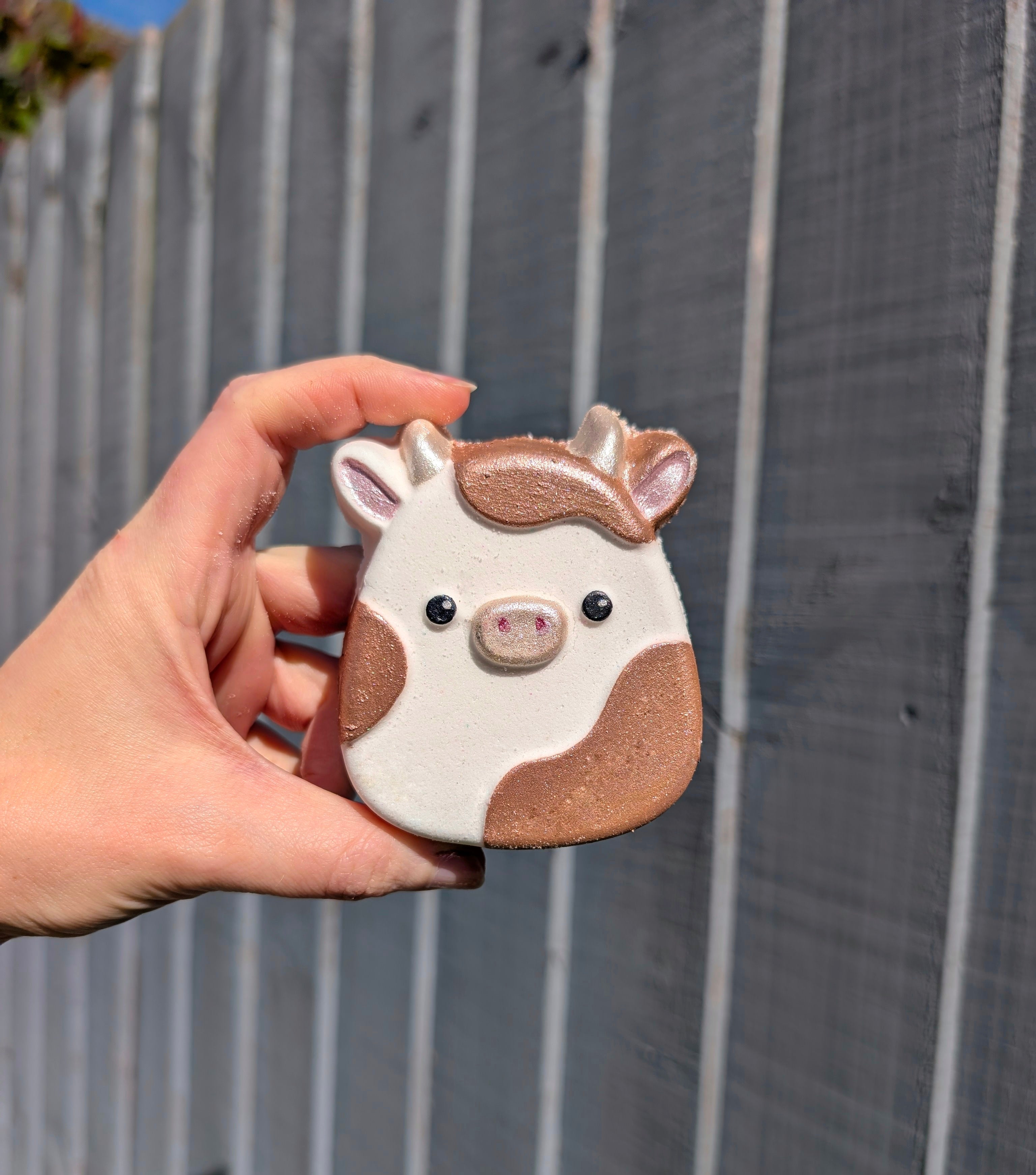 Hand holding a small, cute squishy cow shaped object against a wooden fence background
