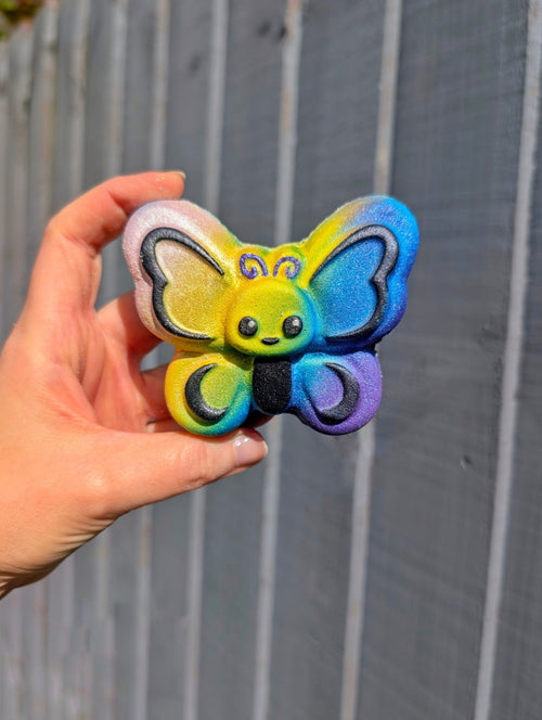 Hand holding a multicoloured butterfly bath bomb, with a grey wooden background.