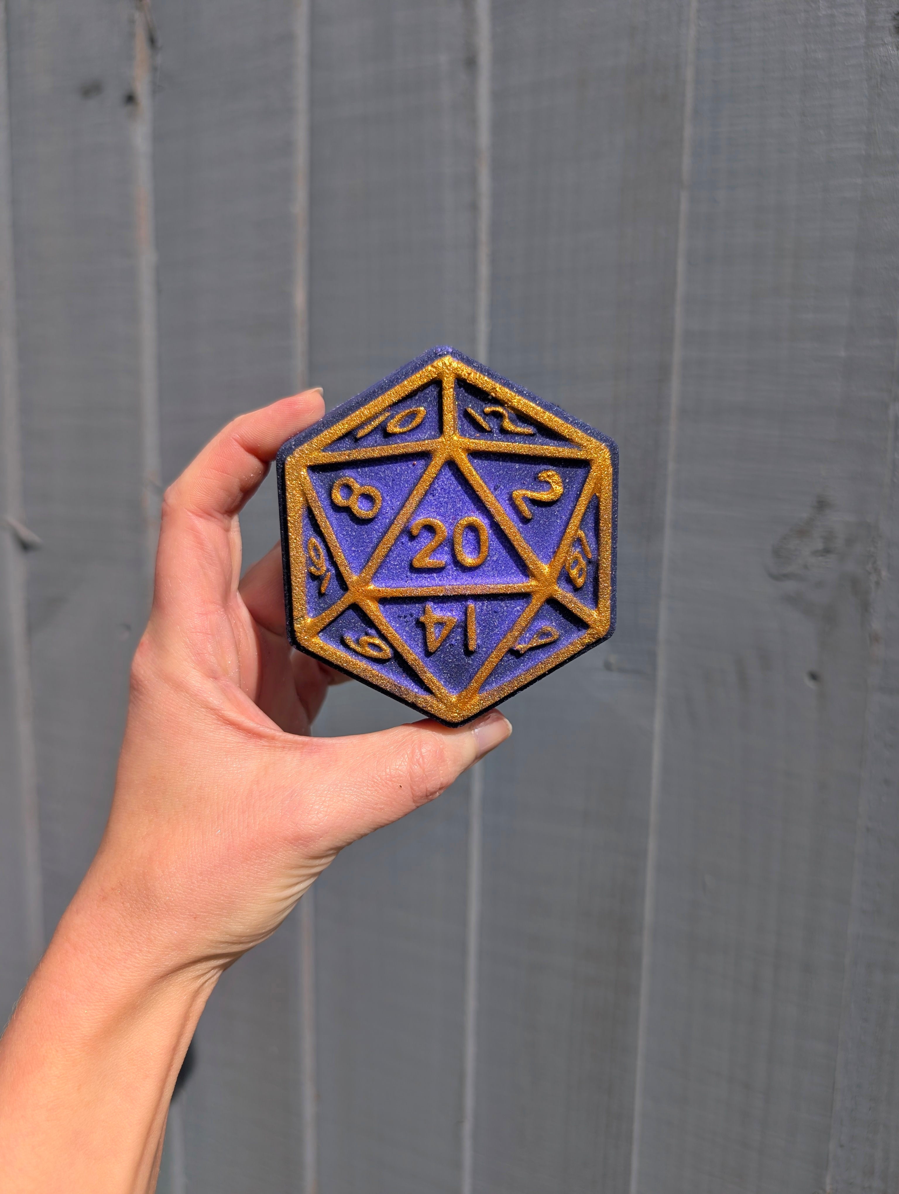 Hand holding a blue and gold D20 dice bath bomb against a wooden background