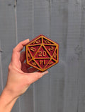 Hand holding a red and gold D20 polyhedral dice bath bomb, against a grey wooden background