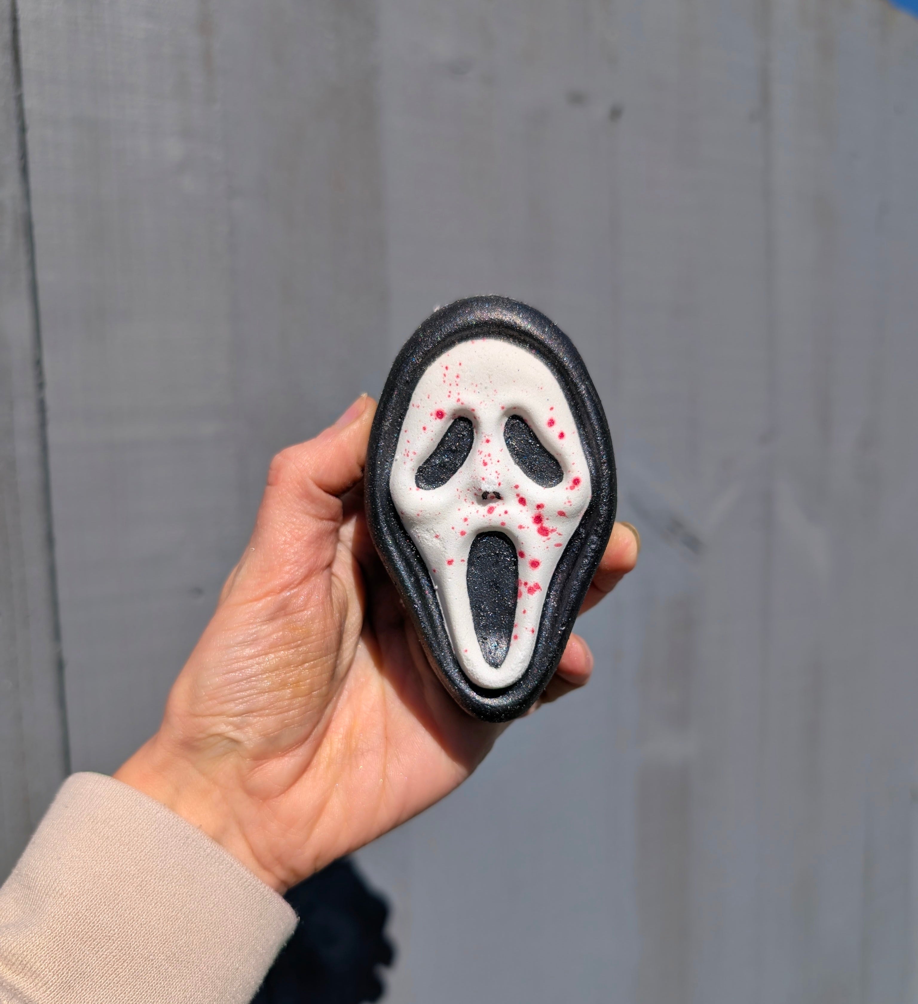 Hand Holding a ghost face from scream shaped bathbomb, on a grey background.