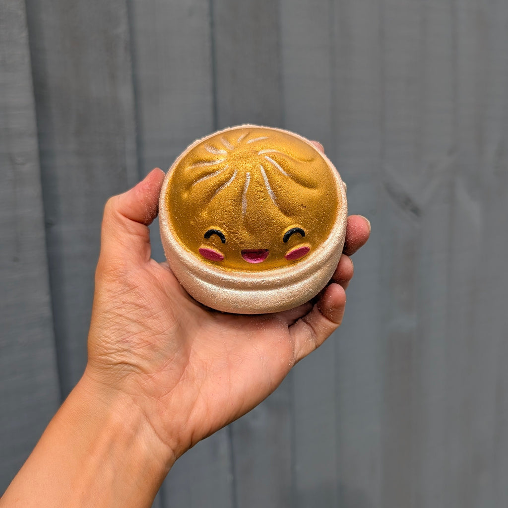 A hand holding a round dumpling bath bomb with a cute smiling face, set against a dark grey wooden plank background.