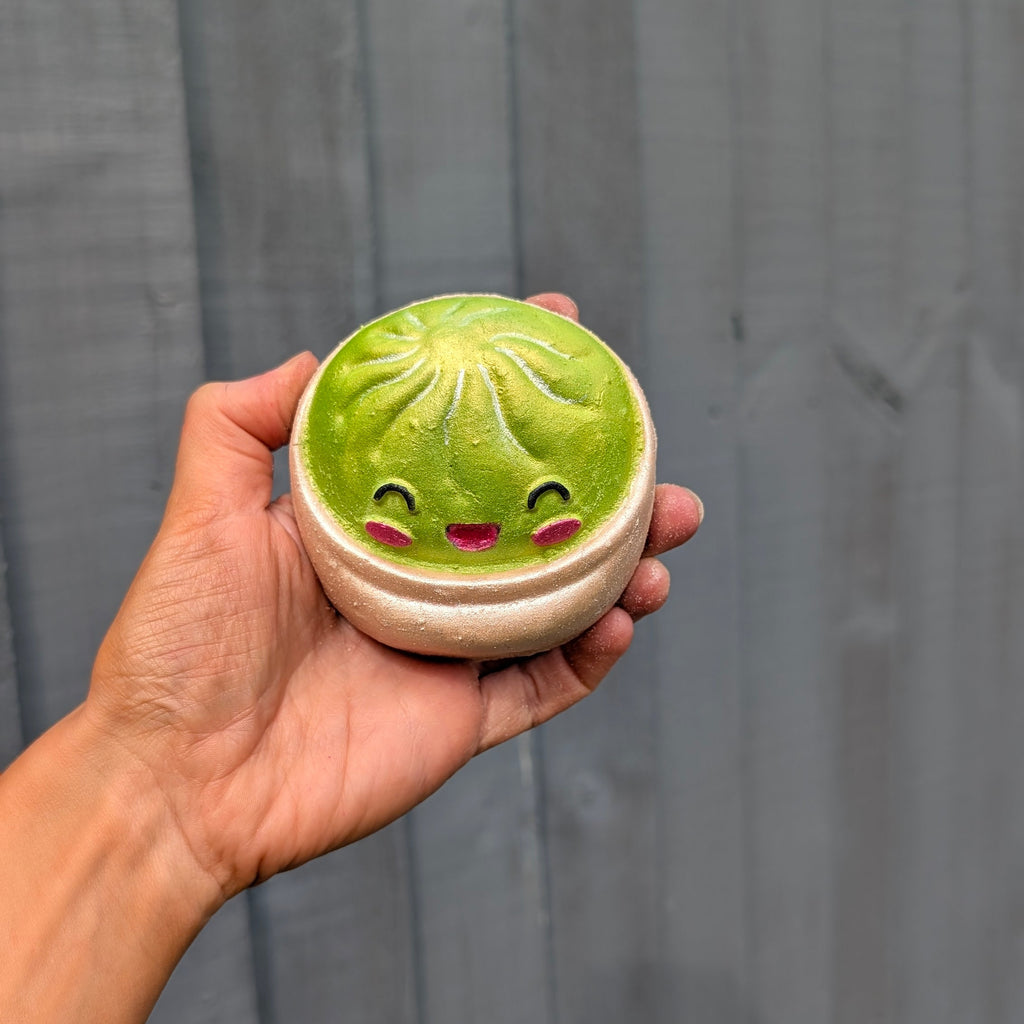 A hand holding a round dumpling bath bomb with a cute smiling face, set against a dark grey wooden plank background.
