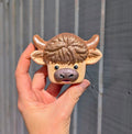 Hand holding a highland cow face shaped bath bomb, with a textured texture against a metal fence background