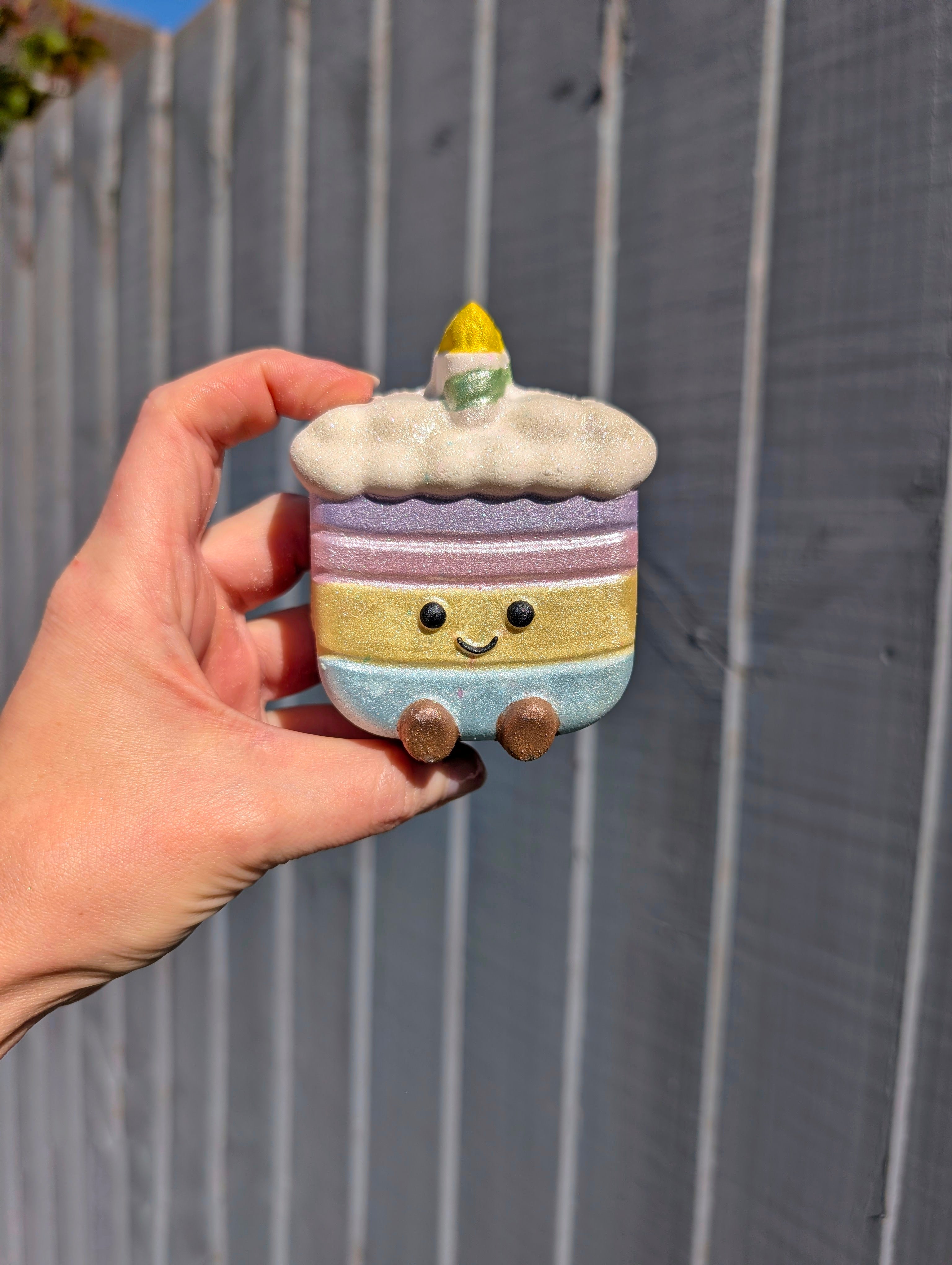 Hand holding a small, colourful, jellycat inspired birthday cake bath bomb, against a wooden fence background.