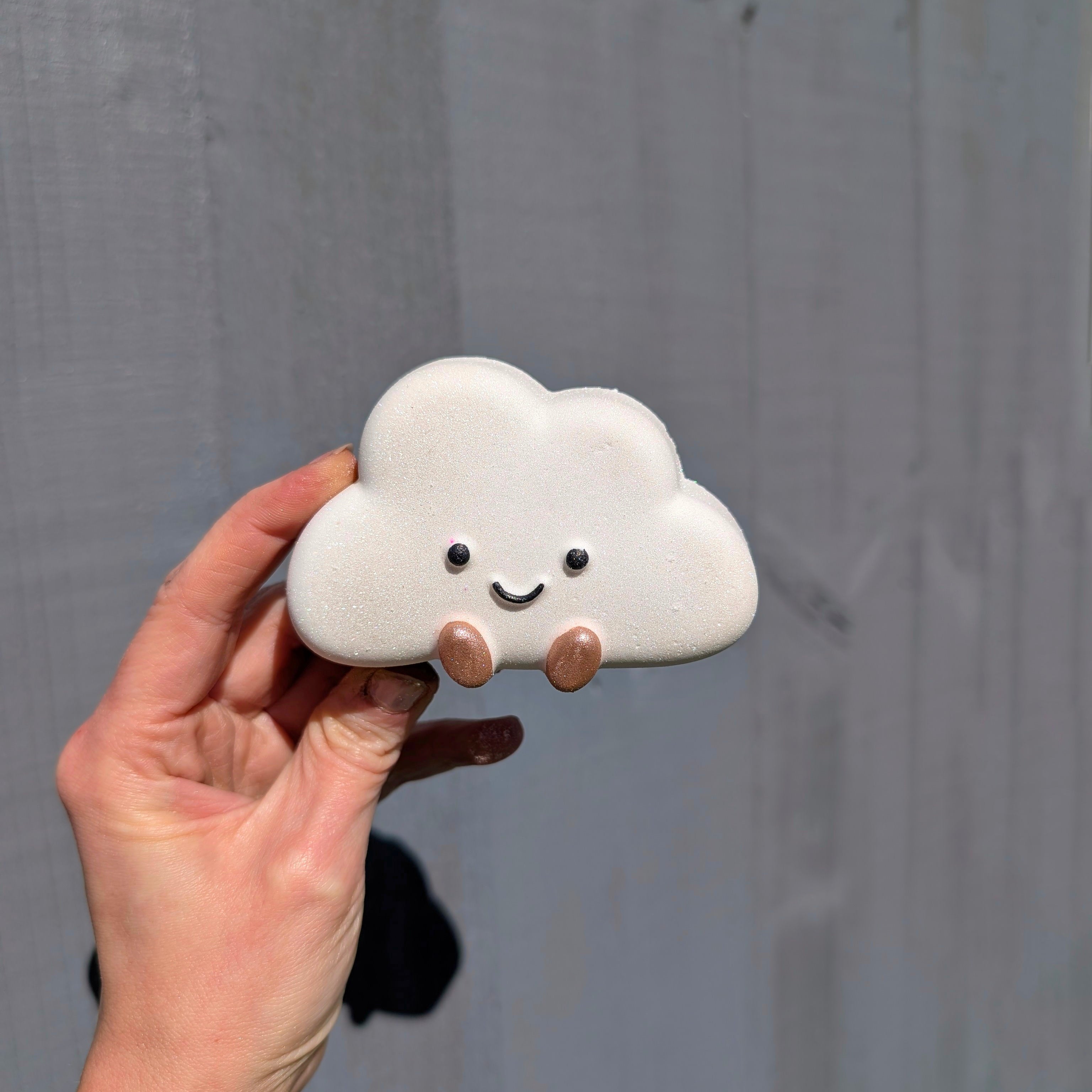A Hand holding a white jellycat cloud shaped bath bomb, on a grey background.