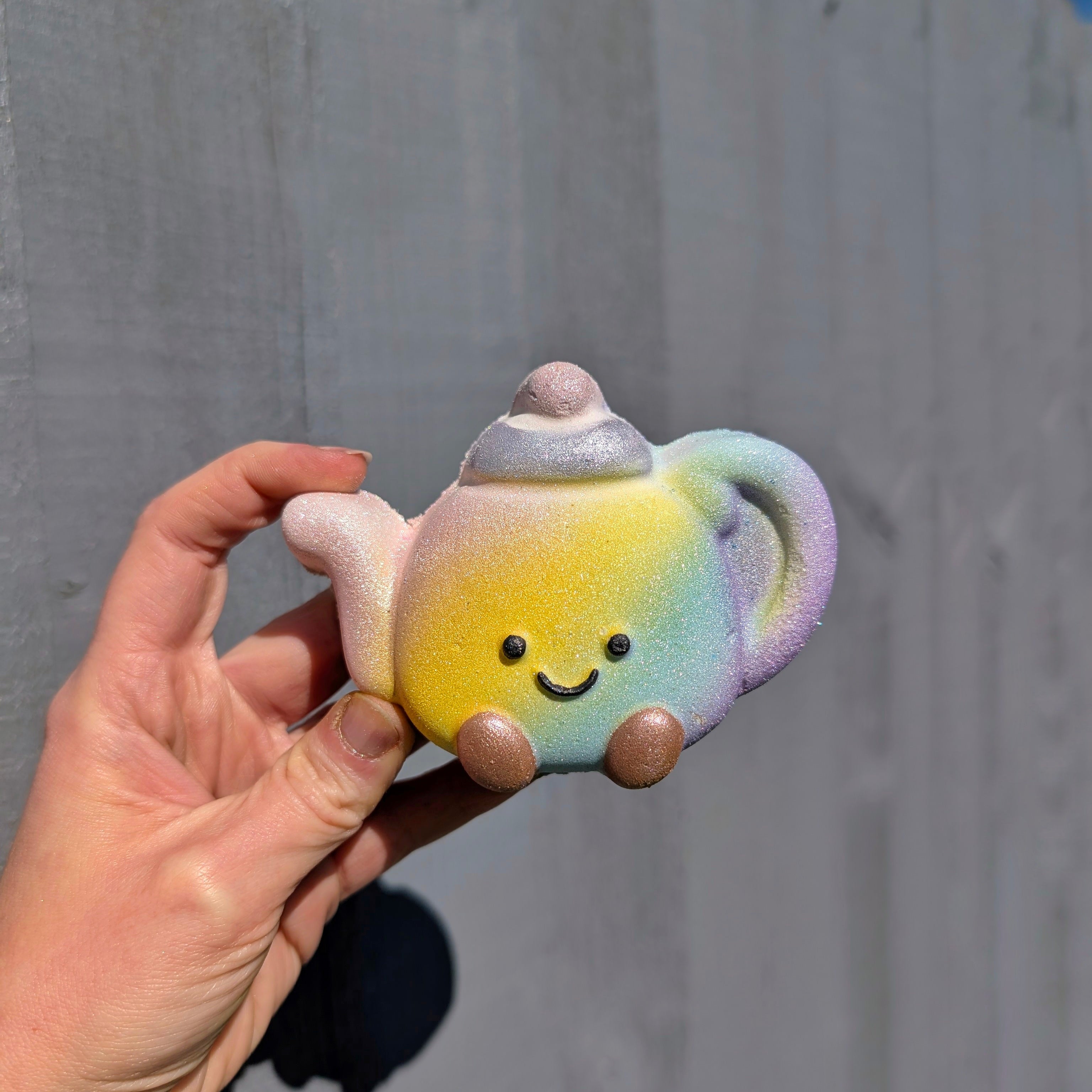 Hand Holding a jellycat teapot shaped bathbomb, on a grey background.
