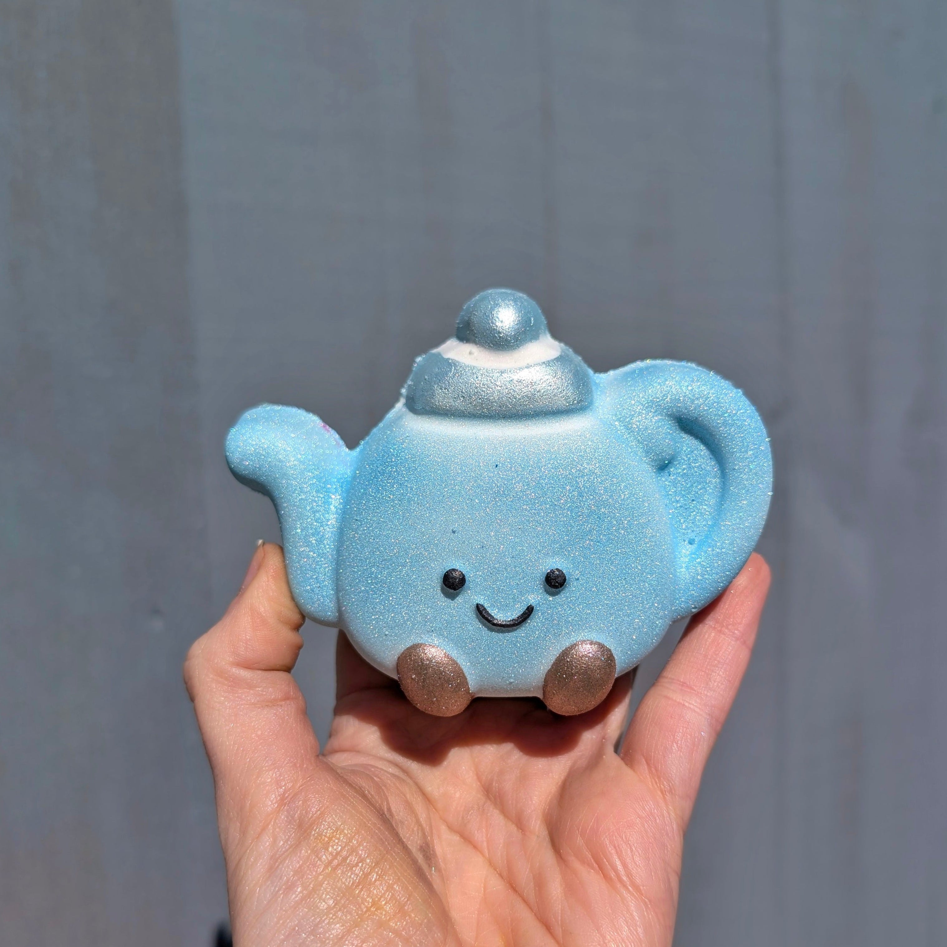 Hand holding a blue bath bomb, shaped like a jellycat teapot, with a grey background.