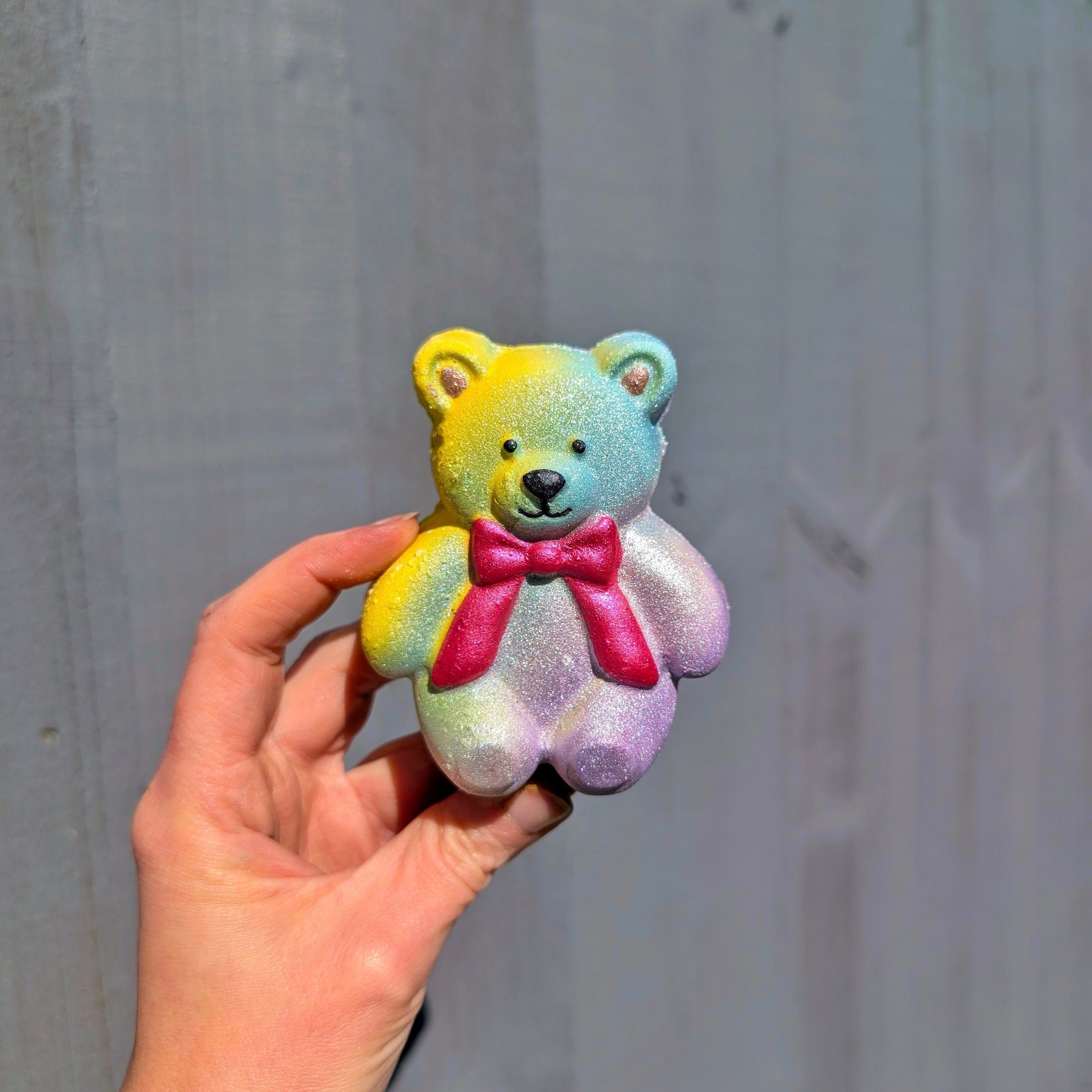 Multi colour jellycat teddy bear shaped bath bomb, held by a hand against a gray wall.