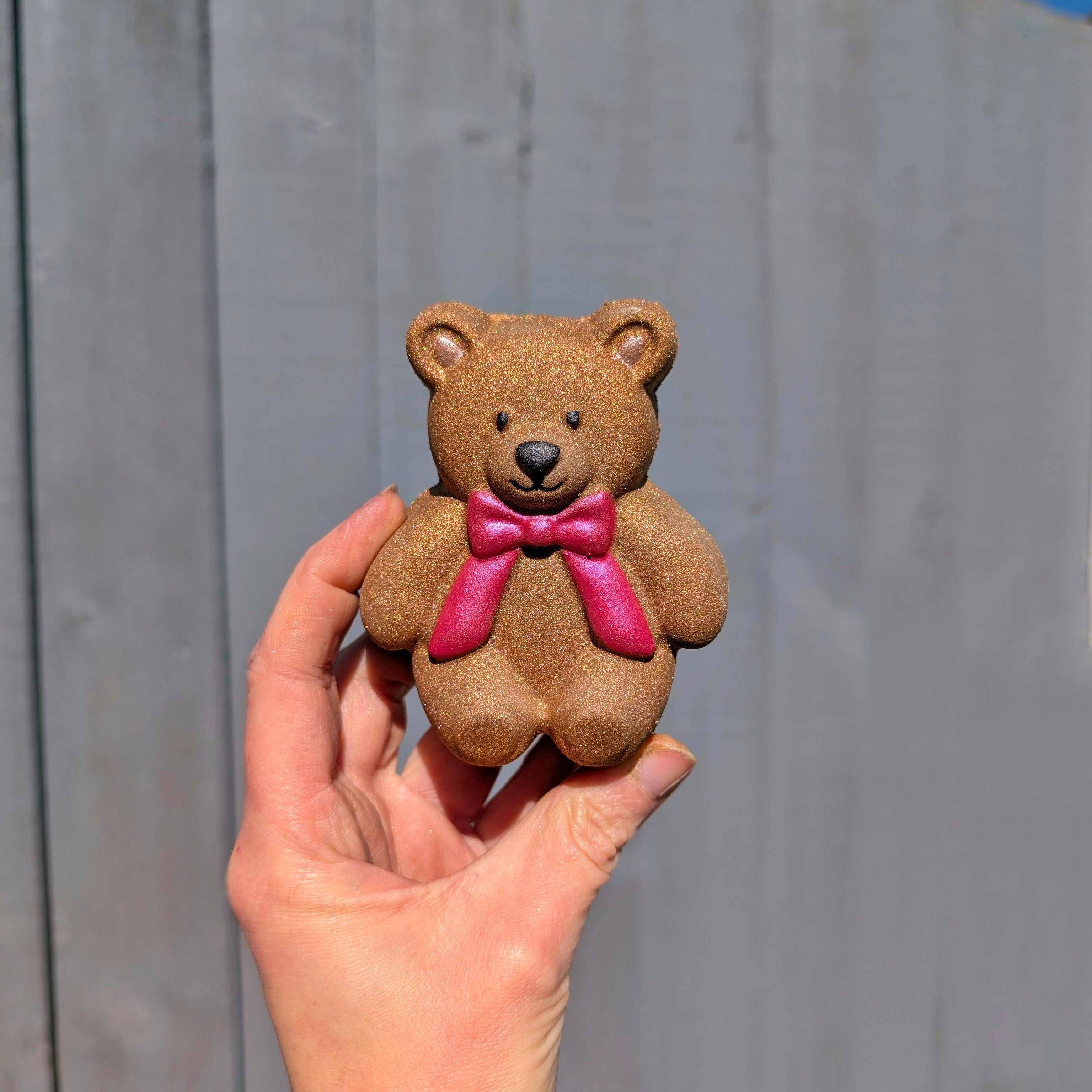 Brown jellycat teddy bear shaped bath bomb, held by a hand against a gray wall.