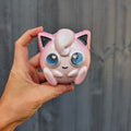 Hand holding a pink jigglypuff shaped bath bomb, against a grey fence.