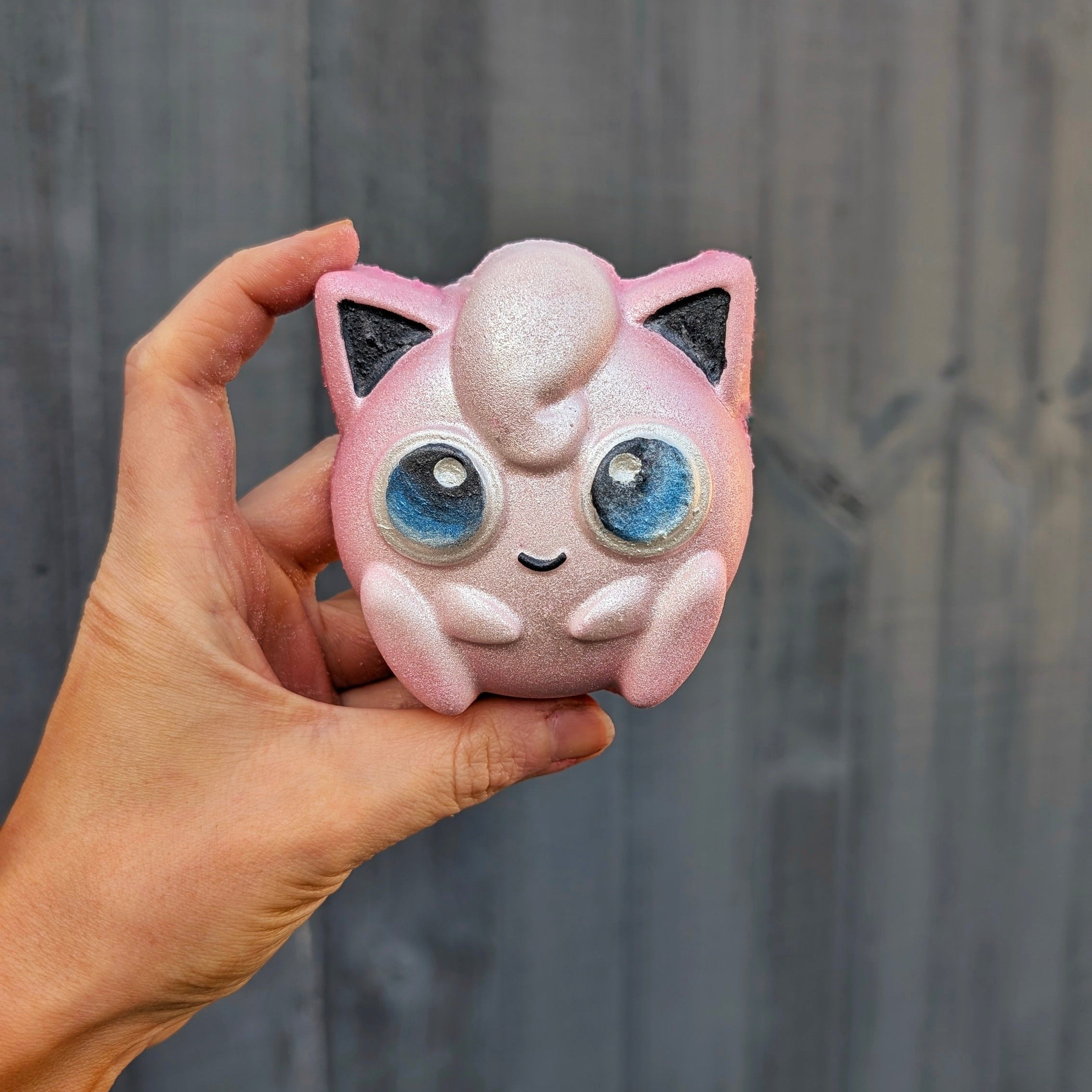Hand holding a pink jigglypuff shaped bath bomb, against a grey fence.