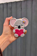 Hand holding a koala-shaped bath bomb with a pink heart and flower against a gray background