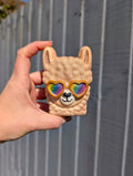 Hand holding a llama-shaped bath bomb with rainbow heart sunglasses against a grey fence background