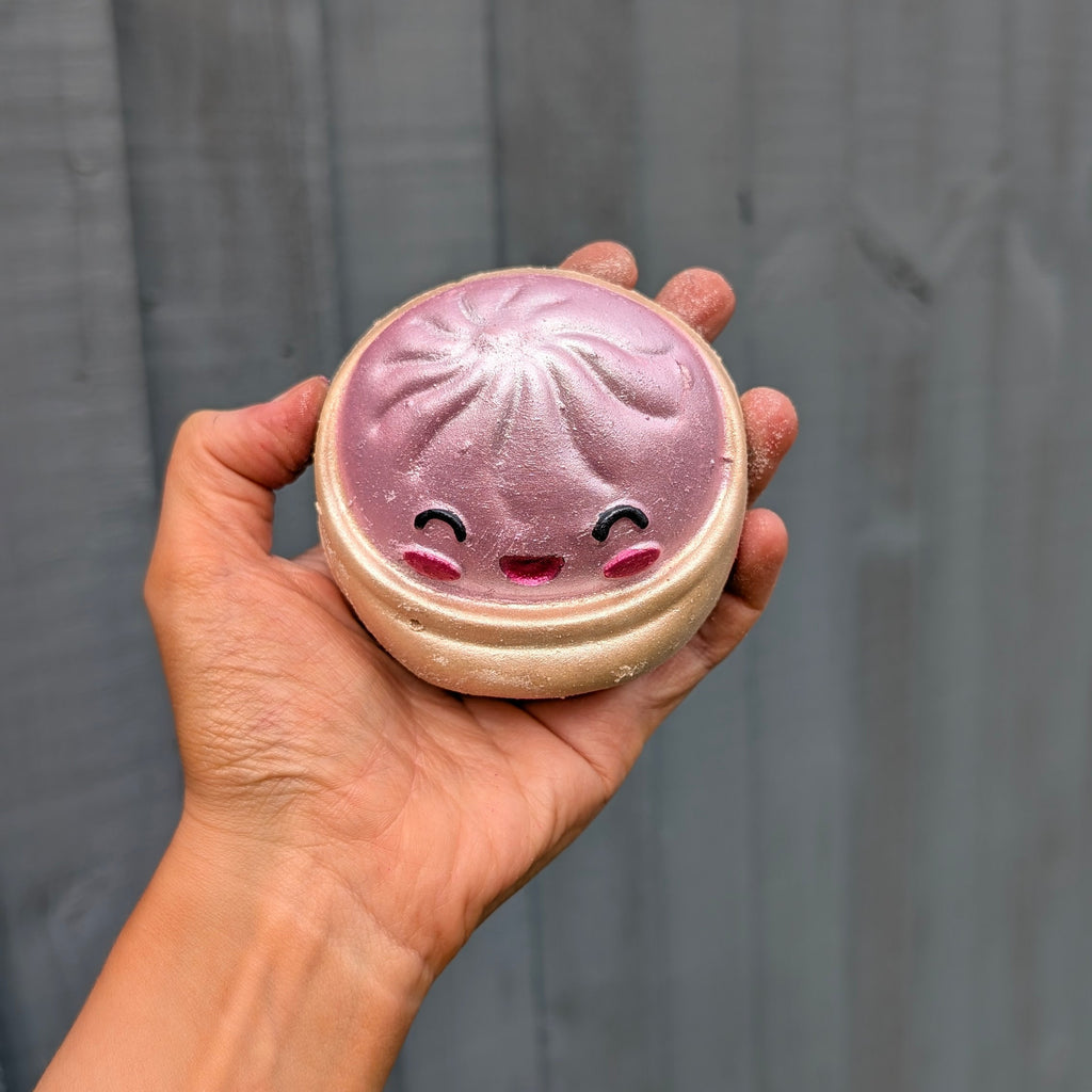 A hand holding a round dumpling bath bomb with a cute smiling face, set against a dark grey wooden plank background.