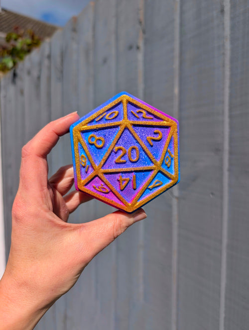 Hand holding a large D20 dice bath bomb with a colourful iridescent finish against a wooden fence background.