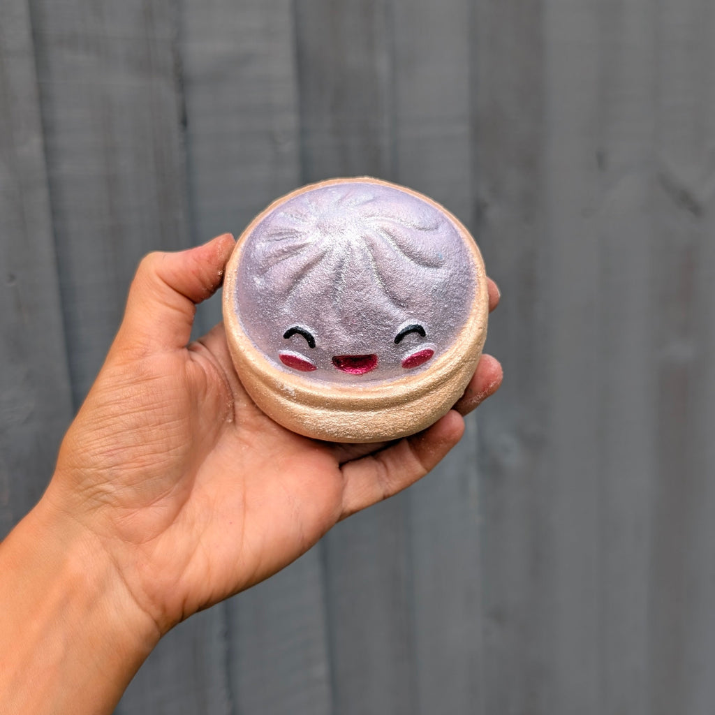 A hand holding a round dumpling bath bomb with a cute smiling face, set against a dark grey wooden plank background.
