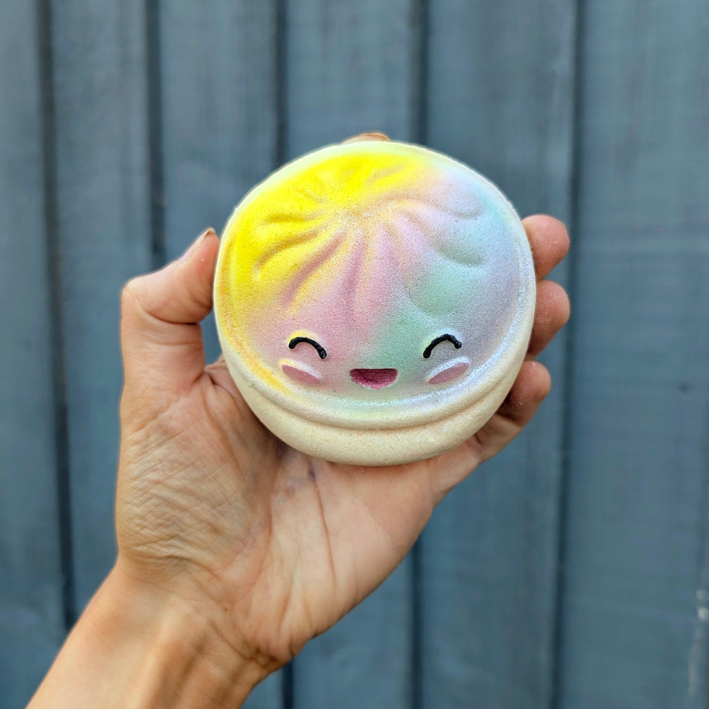 A hand holding a round dumpling bath bomb with a cute smiling face, set against a dark grey wooden plank background.
