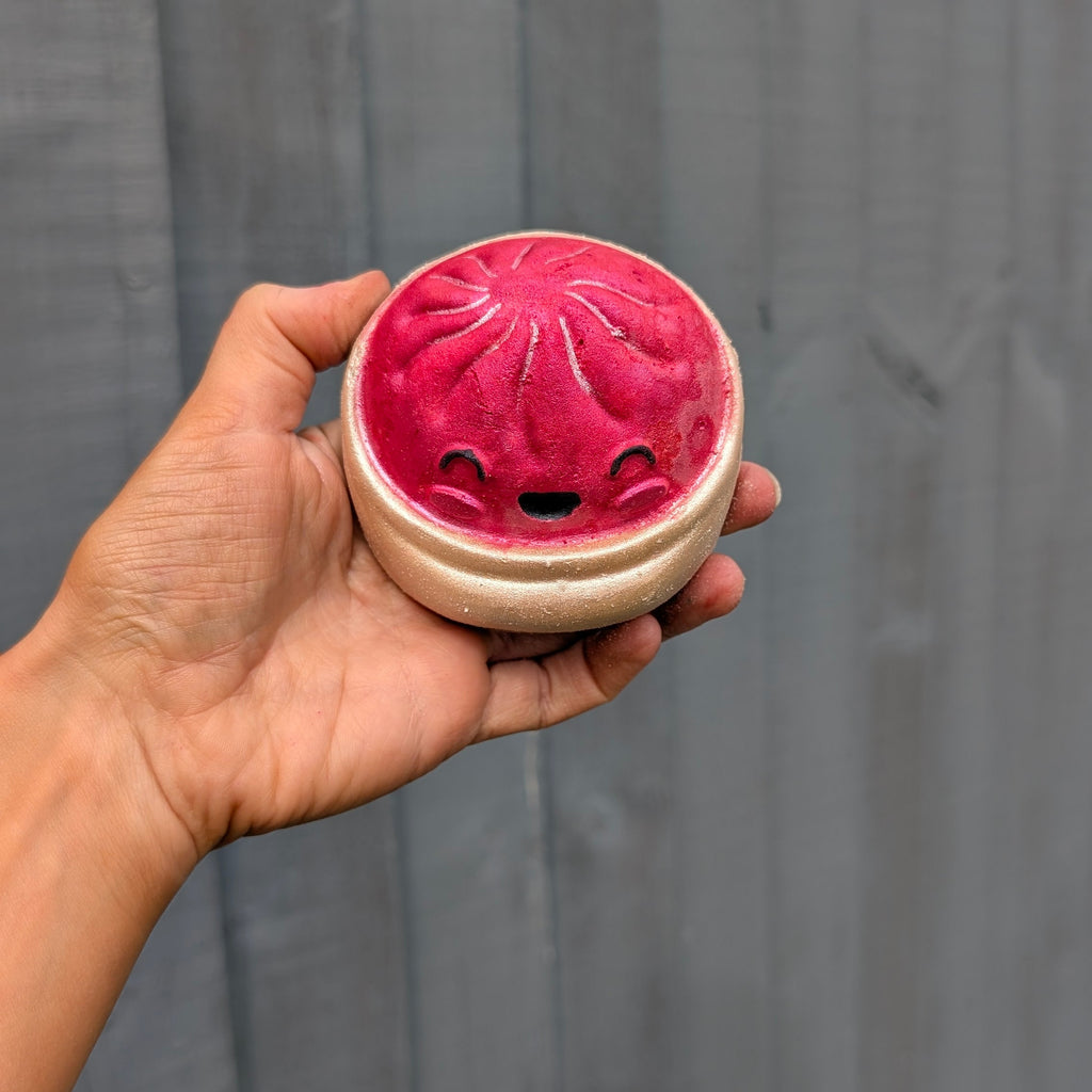 A hand holding a round dumpling bath bomb with a cute smiling face, set against a dark grey wooden plank background.