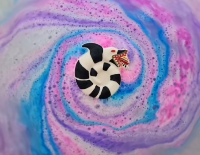 A sandworm shaped bath bomb from Fizz Bomb Cosmetics, fizzing in the bath. Featuring red and black detailing with pink and purple colours in a swirl pattern– a fun, beetlejuice themed bath treat perfect for gifting or relaxing self-care days.