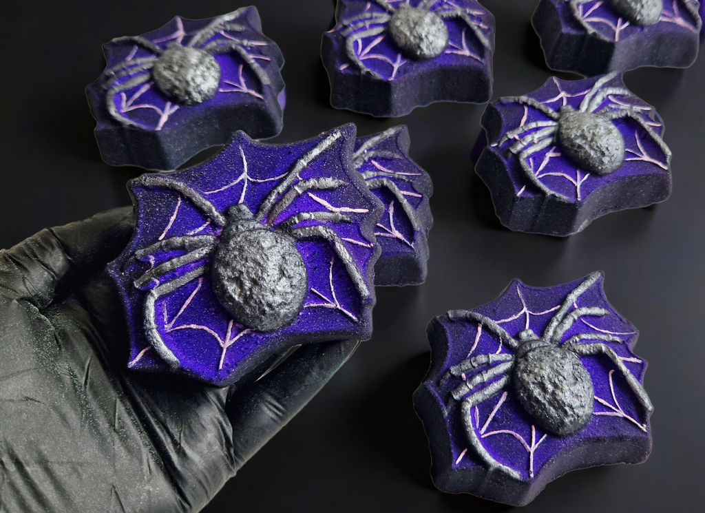 Hand holding a spider in a spiderweb shaped bath bomb from Fizz Bomb Cosmetics, on a black background. Featuring silver and white detailing – a fun, halloween themed bath treat perfect for gifting or relaxing self-care days.