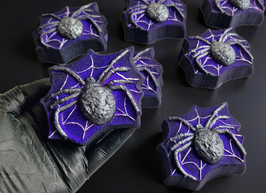 Hand holding a spider in a spiderweb shaped bath bomb from Fizz Bomb Cosmetics, on a black background. Featuring silver and white detailing – a fun, halloween themed bath treat perfect for gifting or relaxing self-care days.